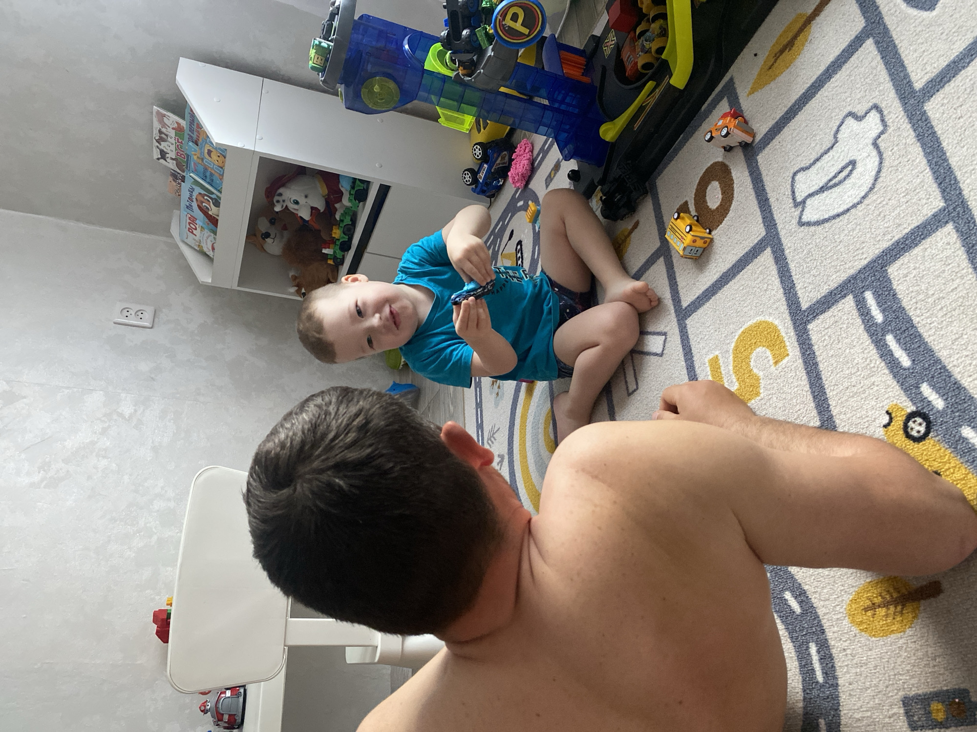 A wide-angle, indoor shot shows a child and an adult on a rug. The child, wearing a blue shirt, is sitting on the rug and holding a toy car. The rug has a pattern resembling streets. The adult is positioned in the foreground, with their back to the camera. They are shirtless. To the left, there is a white chair. Behind the child, there is a toy garage, toys, and a white shelf filled with toys and books. The image appears to be taken at a high angle. There is no evidence to suggest the image was taken in Sochi, Russia.