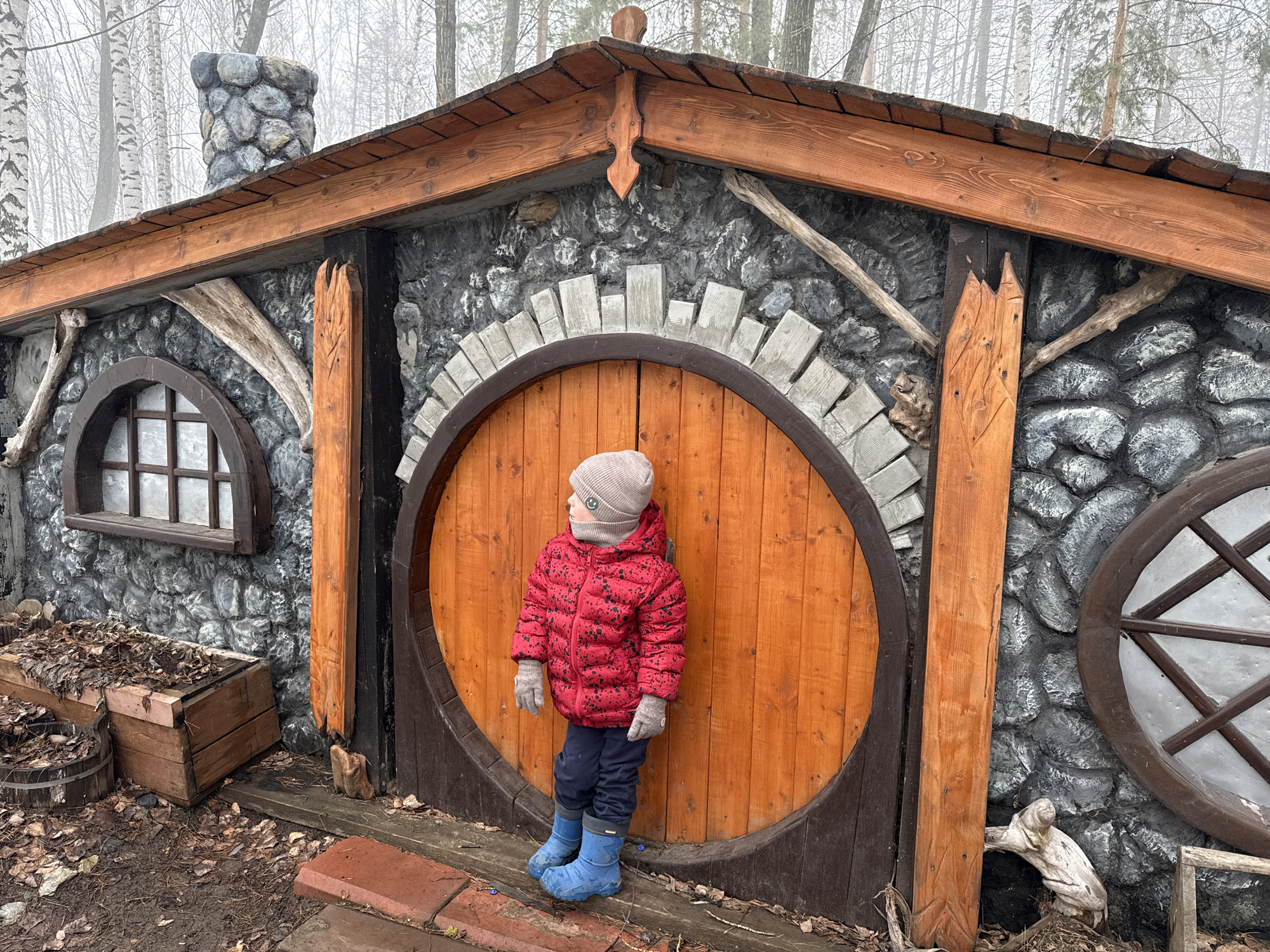 A young child in a red puffy jacket, blue pants, and blue boots stands in front of a whimsical structure. The building has a wooden roof and a grey stone-like wall. The entrance is a large, round wooden door. To the left of the door is a small, arched window, and to the right is a round window. A stone chimney is visible on the left side of the building. The ground is covered in leaves and damp earth. The photo was taken in Chaikovsky, Russia.