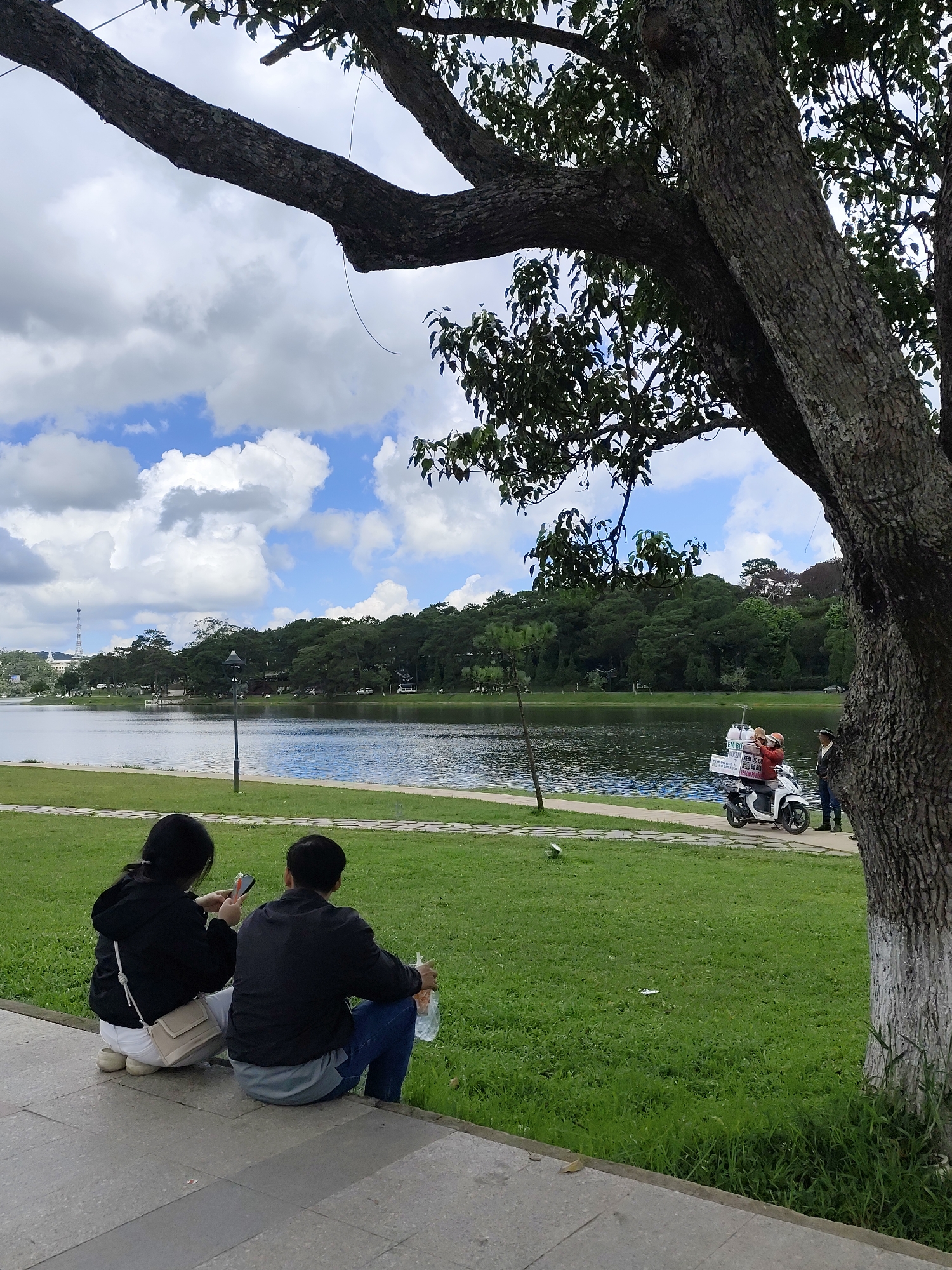 This is a photograph taken in the city of Da Lat, Vietnam, likely at Xuan Huong Lake, a popular tourist destination. In the foreground, two people are seated on a paved walkway, facing away from the camera. The person on the left, wearing a black hoodie and white pants, is holding a smartphone. The person on the right, wearing a black jacket and blue jeans, is holding a plastic bag.
Beyond the walkway is a lush green lawn, and further on, the calm surface of Xuan Huong Lake reflects the sky. The lake is bordered by a dense line of trees, with a road running behind them. In the background, a tall tower can be seen in the distance, and the sky is filled with white, fluffy clouds.
On the path near the lake, a white scooter is parked. A person wearing a red helmet is seated on the scooter, and another person is standing beside it. The scooter is equipped with a carrier on the back, which appears to be holding several items, possibly for sale. The overall scene is serene, with elements of nature and urban life coexisting.
