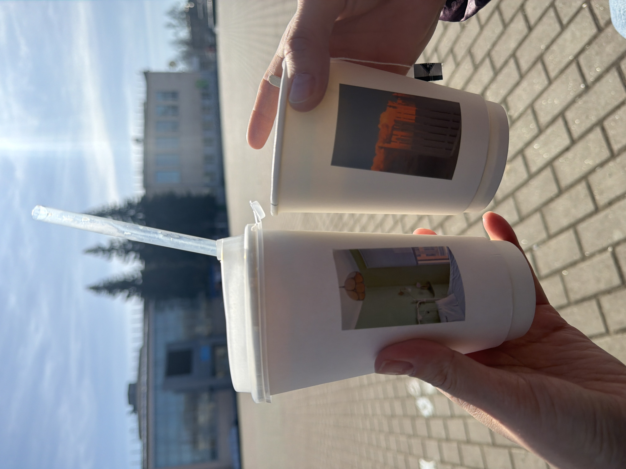 Two white paper cups, each adorned with a photograph, are held in outstretched hands. The top cup has a picture of what appears to be an illuminated building at night, possibly a theater or concert hall. The bottom cup features a photograph of what looks like a tidy hospital room, with a made bed and a lamp. A clear plastic straw protrudes from the lid of the bottom cup. In the background, out of focus, is a city landscape with buildings, trees, and a paved area that suggests a public square or street. The setting appears to be in Chaikovsky, Russia. The presence of the buildings, the paved area, and the cups suggest a casual outing or a break in an urban environment.