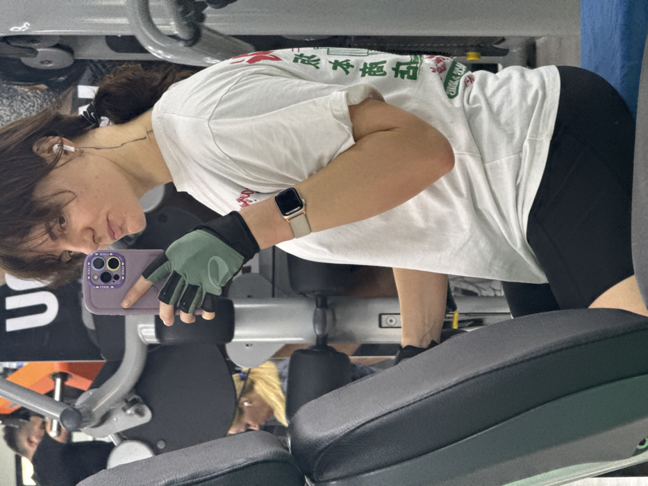 A female subject is depicted seated on a black and grey upholstered gym machine, taking a mirror selfie with a purple-cased smartphone held in her left hand. She has brown hair, partially styled with bangs and pulled back into a ponytail. Her attire consists of a white short-sleeved t-shirt with a graphic design on the back featuring red, green, and black Japanese characters, black athletic shorts, olive green and black fingerless workout gloves, and a beige/rose gold Apple Watch on her left wrist. White wireless earbuds are visible in both ears, with a black cable dangling behind her left ear. Her gaze is directed towards the smartphone camera.
The immediate environment is an indoor gym. In the background, to the subject's left, a man with dark hair in a black t-shirt is partially visible, operating another piece of gym equipment. Further in the background, a blonde-haired female wearing a light-colored top and white earbuds is also seated on gym equipment. Various grey metallic gym machines and black upholstered padding are present throughout the scene. A large white sign with the partial black letters "U" and "M" is visible on a wall in the upper background. Based on the "UM" logo, the location is identified as an Ultimate Montenegro gym, specifically located in the city of Budva, Montenegro.