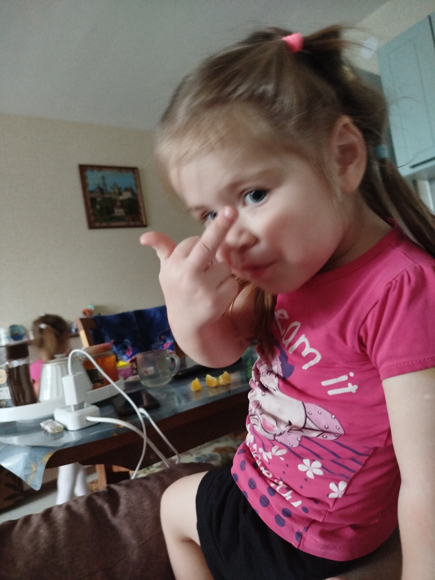 A young girl is sitting on a brown upholstered piece of furniture, wearing a pink t-shirt with a graphic design and black shorts. She is holding up her index finger to her nose, possibly touching or near it. She has light brown hair pulled back in a ponytail with a pink hair tie and has blue eyes.
In the background, on a table covered with a blue and clear patterned covering, there are various items including a white electric appliance, a glass, and cut citrus fruits. Reflected in the table is another child wearing the same pink t-shirt.
The background also includes a framed picture hanging on a cream-colored wall and a glimpse of turquoise cabinets. The photo was taken indoors in what appears to be a private home. The exact location in the city of Chaikovsky, Russia, cannot be determined from this photo.