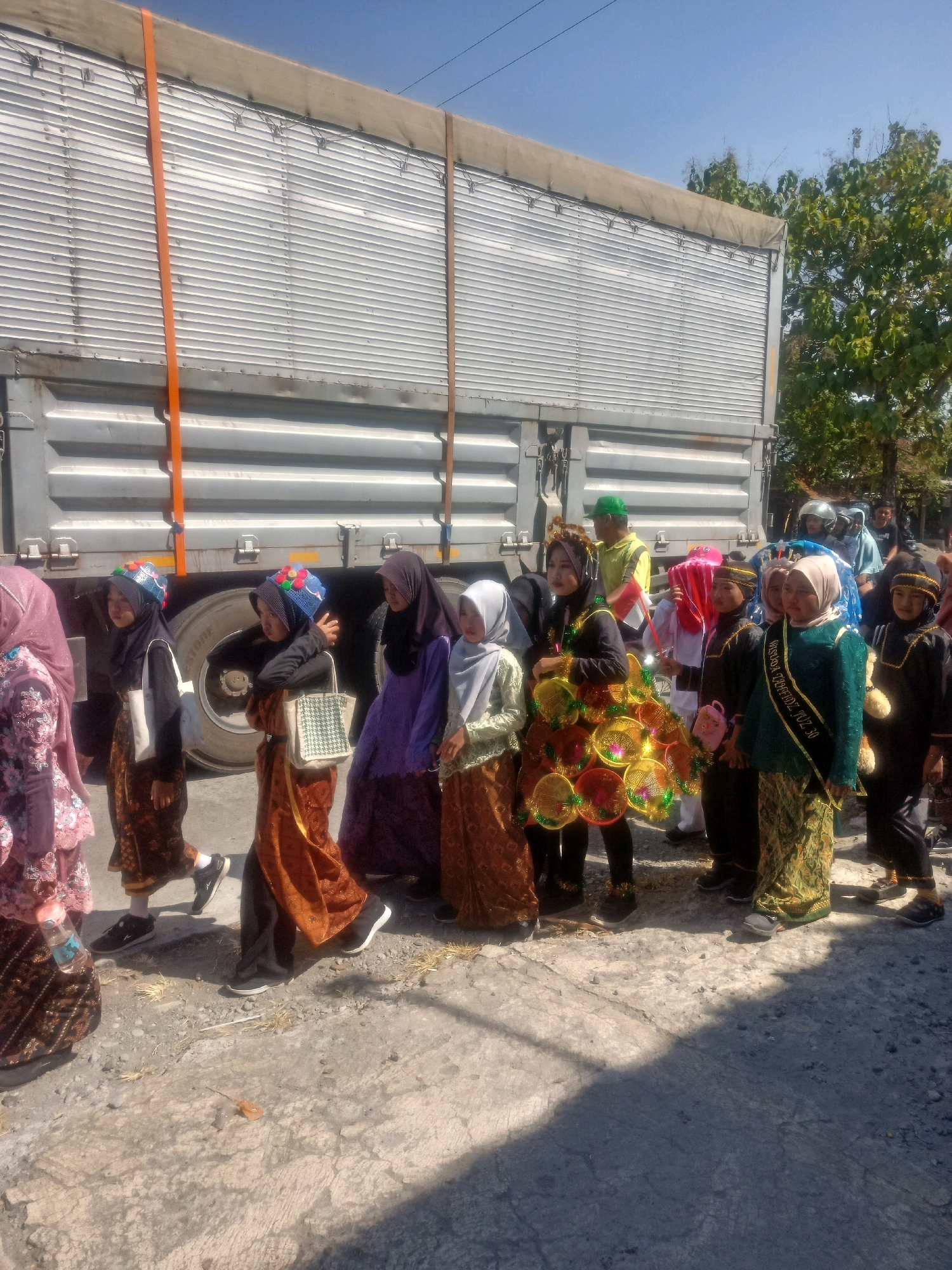 A group of children, mostly girls wearing hijabs and traditional Indonesian attire, are walking in a procession next to a large truck. The truck is silver and appears to be a flatbed with a tarp covering the cargo area. It is secured with orange and brown straps. The children are dressed in various colors, with some wearing elaborate headpieces and sashes. One child in the foreground holds a decorative fan-like object. The ground is paved with concrete and dirt, with some dry grass and debris visible. The sky is clear and blue, and there are trees in the background. The scene appears to be taking place outdoors, possibly during a cultural event or parade in Sumberlawang, Indonesia.