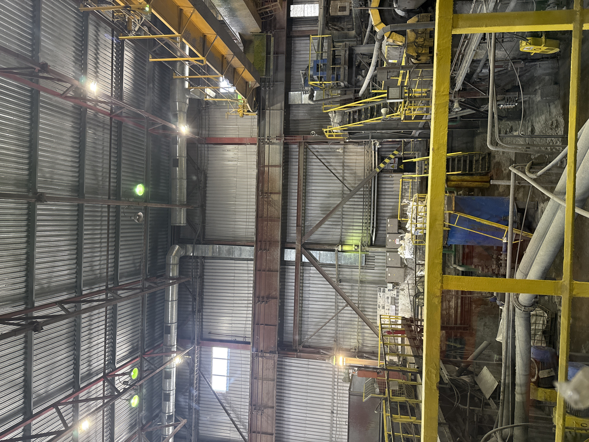 The image shows the interior of a large industrial facility with a corrugated metal roof and walls. The space is filled with various machinery, pipes, and metal structures, suggesting it is a processing or manufacturing plant. Yellow railings and stairs are prominent, indicating elevated walkways and platforms. There are also large ducts and ventilation systems visible. The lighting appears to be a mix of overhead industrial lights and some natural light filtering through windows. The overall impression is one of a busy, functional industrial environment. The location is identified as KOKENTAU RURAL DISTRICT, KAZAKHSTAN.