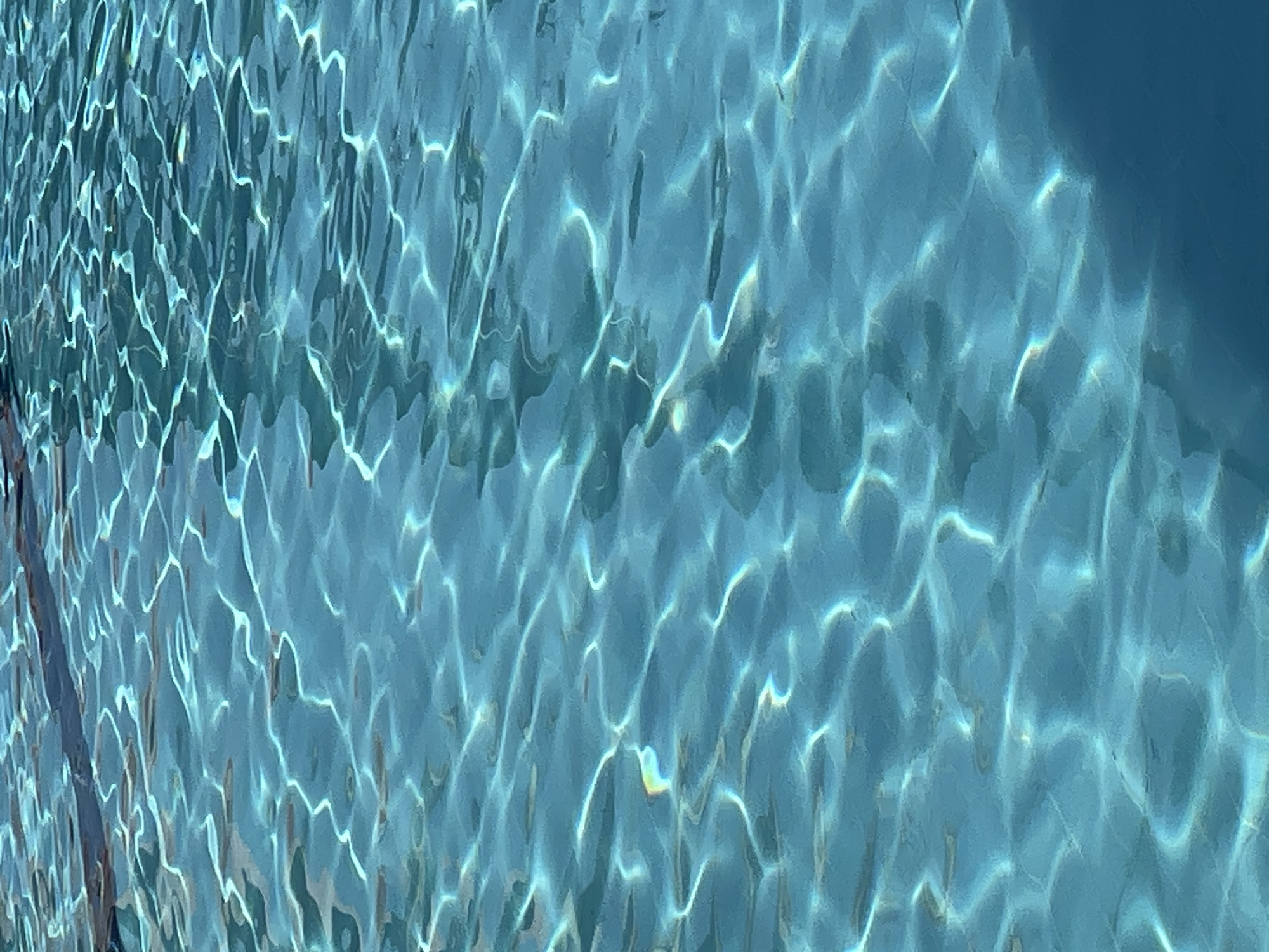 The image shows a close-up view of the surface of a swimming pool in Kuta Selatan, Indonesia. The water is a clear, vibrant blue, with sunlight reflecting off its rippling surface in shimmering patterns of white light. The light creates a dynamic, abstract mosaic across the water, with areas of shadow and bright highlights. There are no people or objects visible in the frame, only the natural movement of the water and the play of light upon it. The overall impression is one of tranquility and the simple beauty of water.