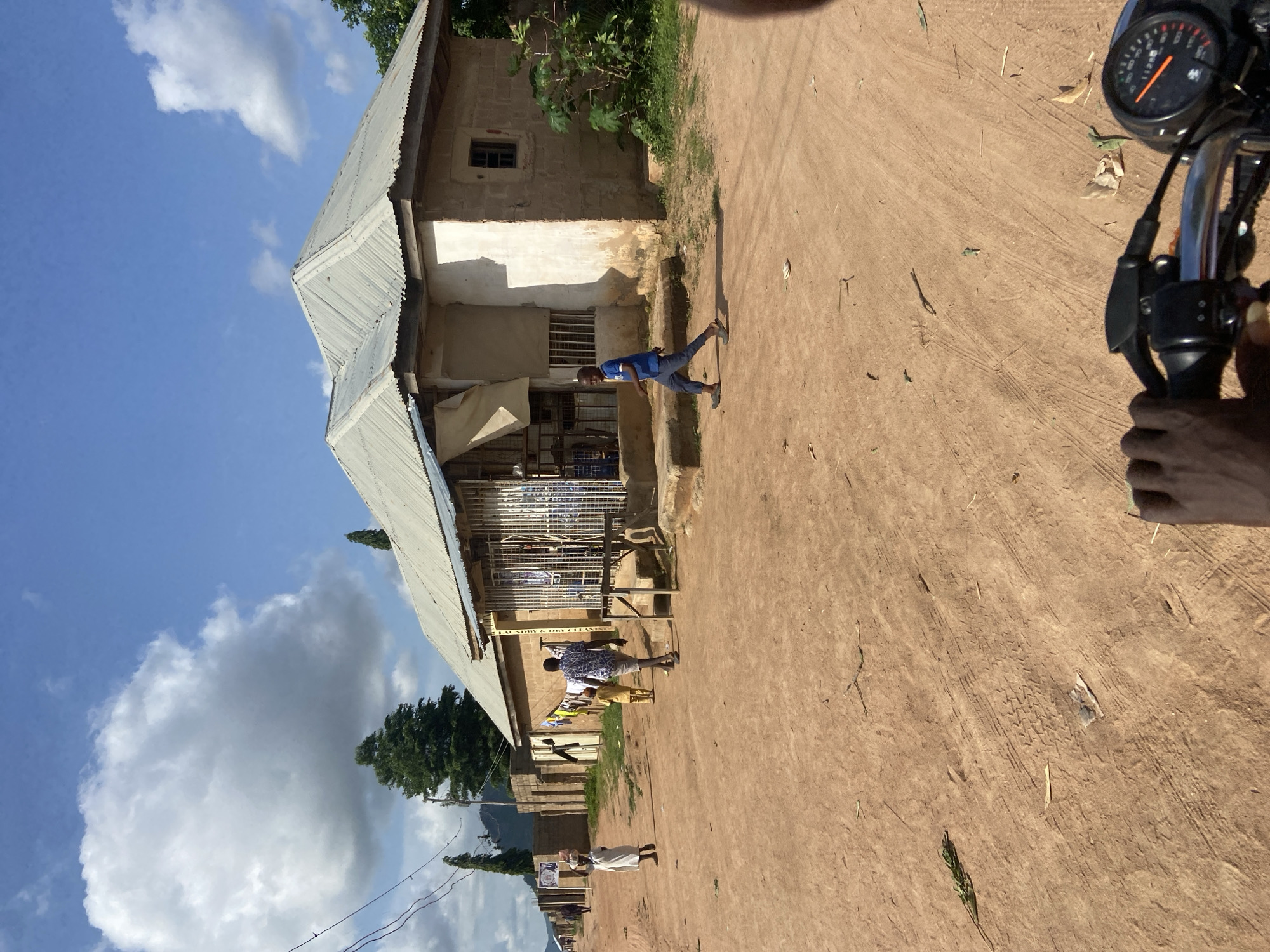 The image depicts a street scene in Kuson, Nigeria. A dirt road runs through the frame, lined with buildings. On the right, a building with a corrugated metal roof and stone walls appears to be a shop or dwelling, with bars on the windows and an open entrance. To the left, a row of similar buildings extends into the background. Several people are visible: a young boy in a blue shirt and dark pants walks across the road in front of the shop, a man in a patterned shirt walks away from the camera, and a woman in a white dress with a basket on her head walks towards the left side of the frame. Other figures are further down the road. The environment is sunny, with a blue sky and scattered clouds. Vegetation is present in the background, including trees. In the foreground, the handlebars and dashboard of a motorcycle are visible, suggesting the photo was taken from a moving motorcycle.