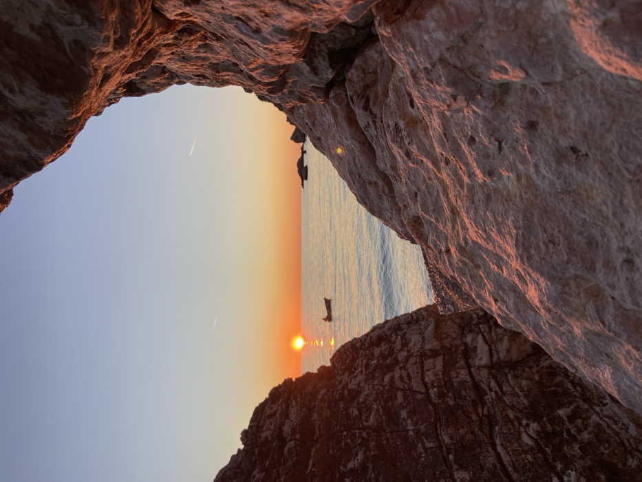 The image depicts a sunset over a body of water, viewed through a natural aperture formed by two large, textured rock formations. The scene is located at an unknown coastal area. The sun, a bright orange disc, is positioned centrally on the horizon, casting a distinct vertical reflection path on the calm water.
A single, small, dark-colored boat is present on the water, slightly to the left of the sun's reflection. A solitary individual is visible in silhouette within the boat, seemingly stationary. Further right on the horizon, several dark, indistinct rock formations or islets are silhouetted against the sky.
The sky transitions from a light blue at the zenith to vibrant orange, yellow, and red hues near the horizon, indicative of a sunset. A thin, white aircraft contrail is visible in the upper-left quadrant of the sky.
The two prominent rock formations in the foreground frame the central view. The left formation is dark brown and heavily textured with deep crevices. The right formation is lighter, appearing beige or light grey, also displaying rough texture and crevices. Both formations are illuminated by the setting sun, casting strong reddish-orange light onto their inner surfaces and contours. The water surface shows minimal ripples.