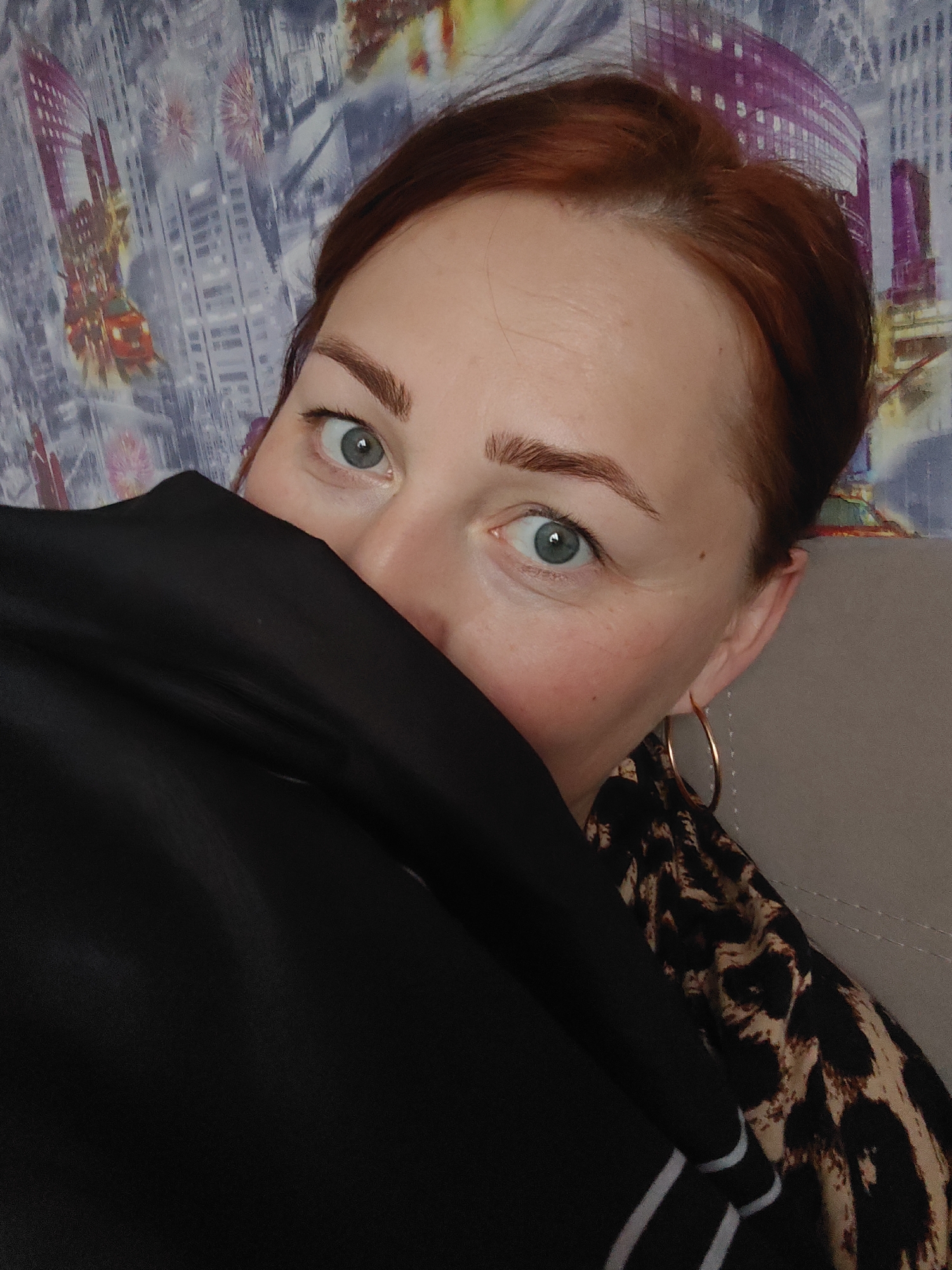 A medium shot depicts a fair-skinned woman with auburn hair pulled back, partially covering her face with a black garment. Her eyes are light blue. She wears a gold hoop earring on her right ear. A leopard-print patterned garment is visible at her shoulder. Behind her is a wall with a patterned design resembling a cityscape with purples, grays, and white tones. The photograph was taken in Markovskii, Russia.