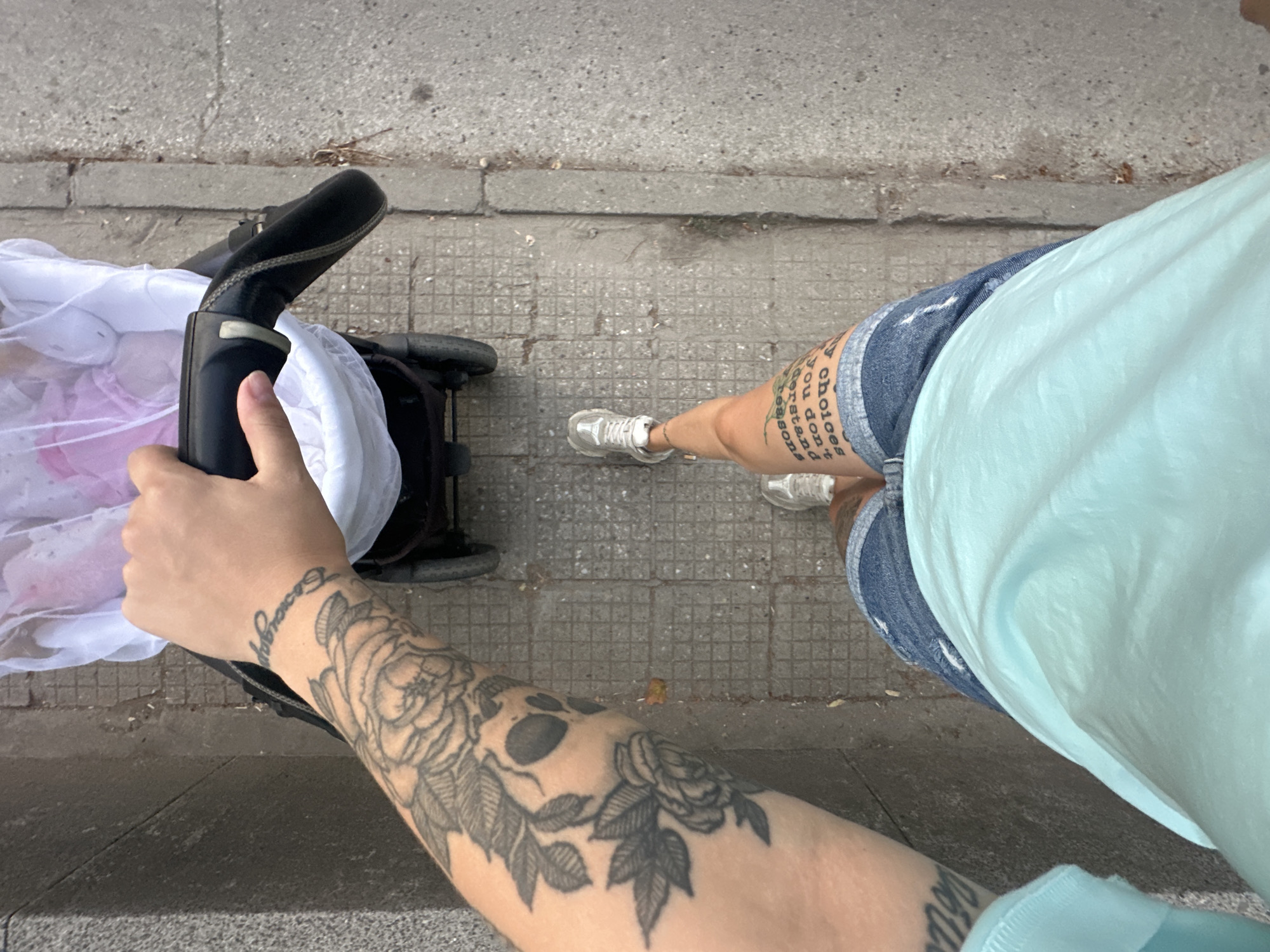 The image is a first-person perspective of someone walking down a sidewalk in the city of Plovdiv, Bulgaria, pushing a baby stroller. The person has tattoos on their left arm and right thigh. The stroller, covered with a white mosquito net, contains a baby. The sidewalk is paved with square tiles and is bordered by a concrete curb and a gray textured wall. The ground appears wet in places, suggesting recent rain. The person is wearing denim shorts and a light blue t-shirt. The lighting indicates it is daytime, with overcast conditions.