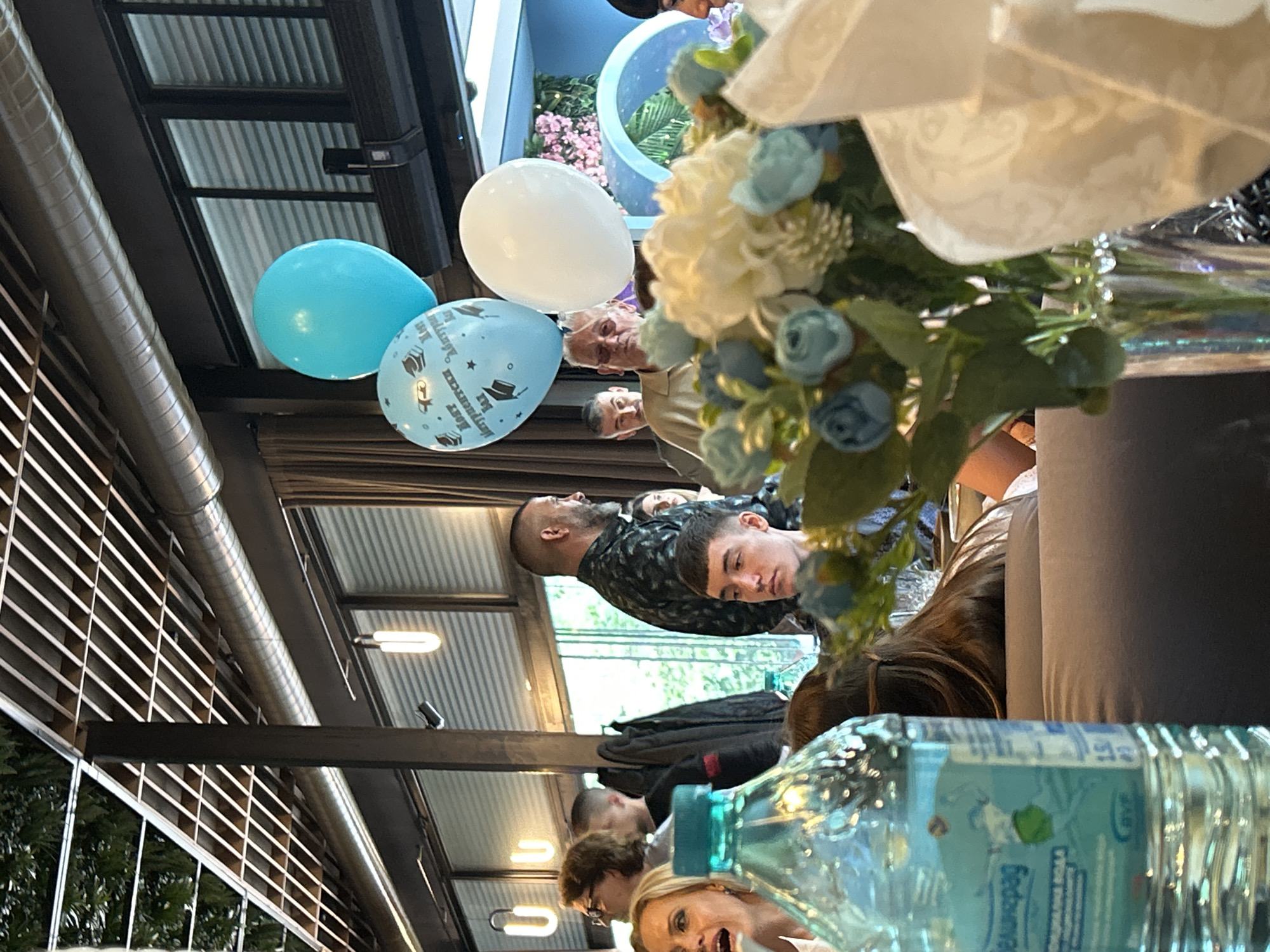 This image shows an interior scene at what seems to be a celebratory gathering in Burgas, Bulgaria, as suggested by the bottle label. The foreground contains a half-empty plastic bottle of water and a floral arrangement with blue and white artificial flowers, which is out of focus. In the mid-ground, several individuals, including a young man looking directly at the camera and other adults, are seated at a table. In the background are balloons, one of which has a graduation cap design. The location appears to be a restaurant or event venue, characterized by a louvered ceiling and lighting fixtures. There is also what seems to be a patio area, visible through the windows, with flowering plants.