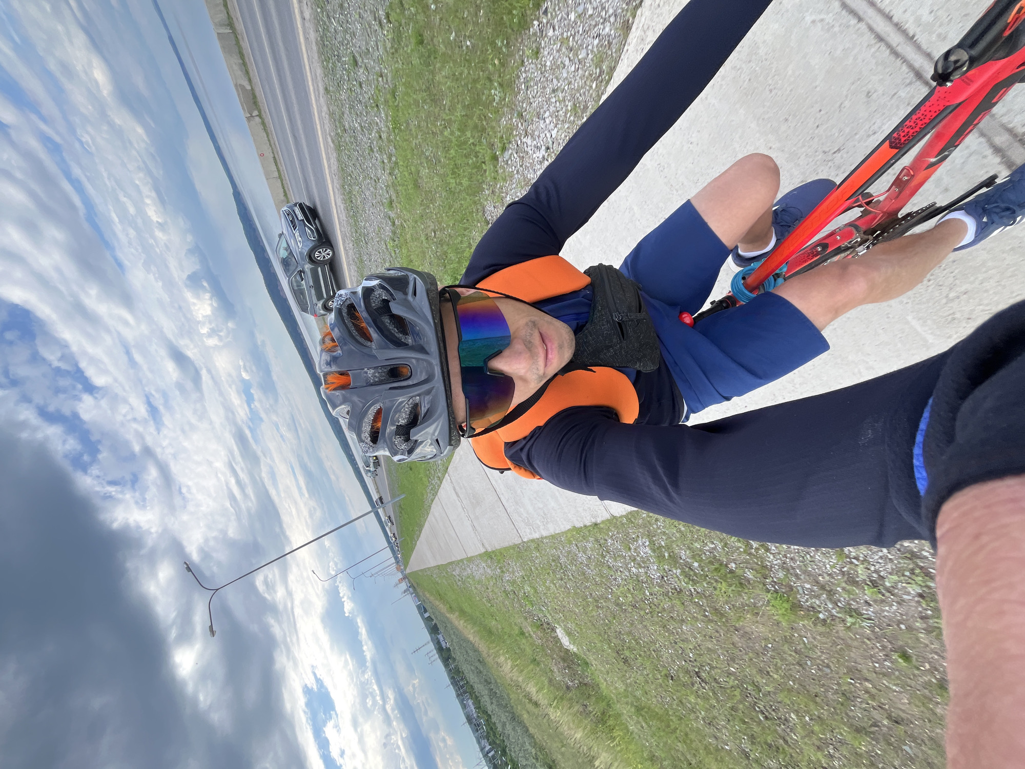 A man rides a red bicycle on a paved path alongside a road in Chaikovsky, Russia. He wears a dark blue long-sleeved shirt, blue shorts, a gray helmet, and mirrored cycling glasses. An orange vest is worn over his shirt, and a black fanny pack is secured around his waist. The road to his right features several cars, and beyond the road is a large body of water. The sky is partly cloudy with a mix of gray and white clouds. Lampposts line the path. The vegetation to his left is grassy and sparsely populated with small plants. The perspective suggests the photo was taken by the rider himself.