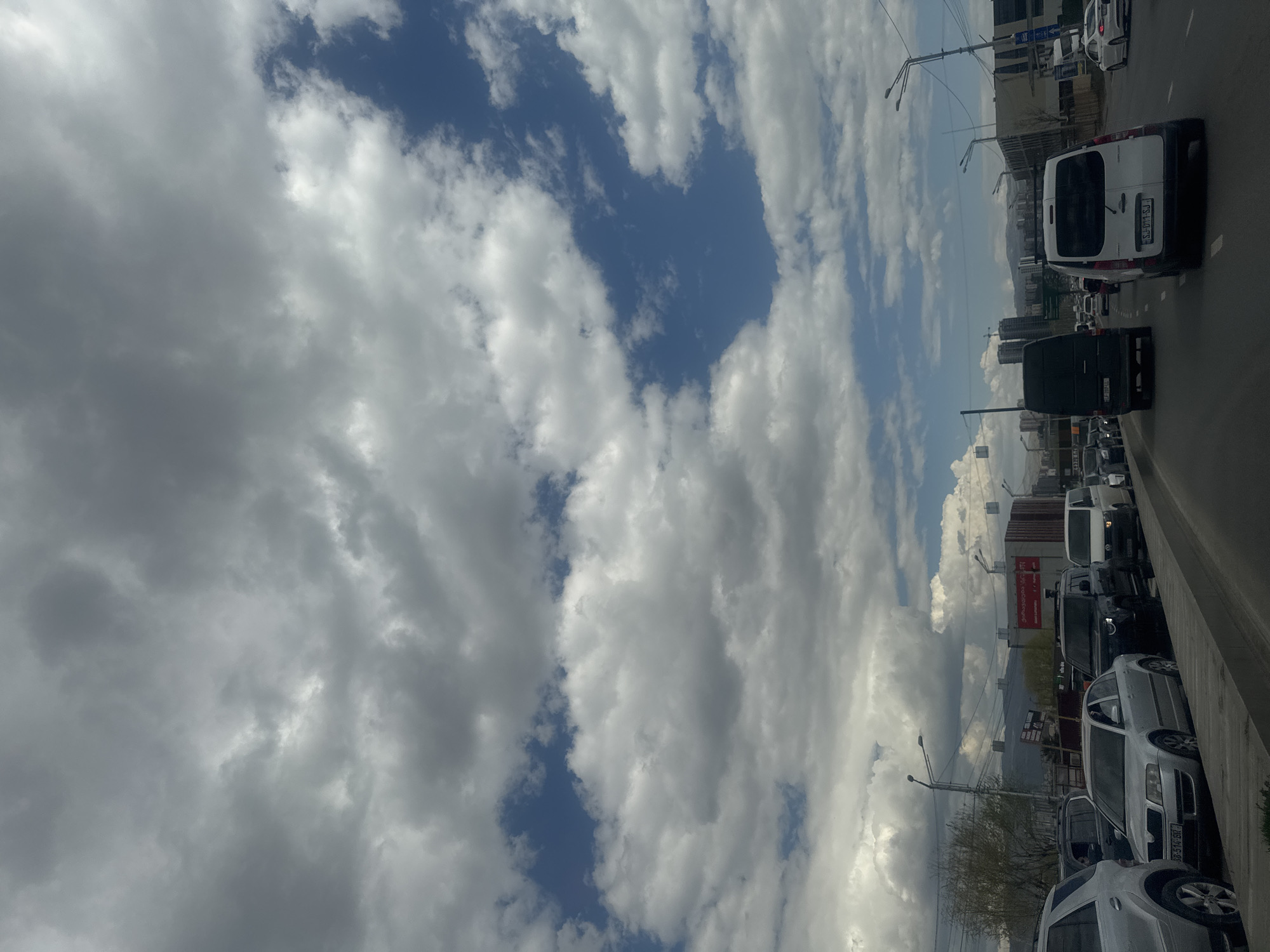 The image is a medium shot of a traffic jam in Tbilisi, Georgia, taken from a slightly elevated perspective. The view is mainly oriented towards the sky, filled with a mix of dense grey and white clouds, interspersed with patches of blue sky.
The lower right of the image reveals a line of cars in a heavy traffic situation. The vehicles are a mixture of vans, sedans, and SUVs, mostly in shades of white, silver, and black. The cars are closely packed together, indicating the congestion.
In the background, there are buildings of various heights, suggesting an urban setting. A few utility poles and streetlights are visible. The overall atmosphere suggests a busy day with overcast weather in a city that experiences frequent traffic congestion.