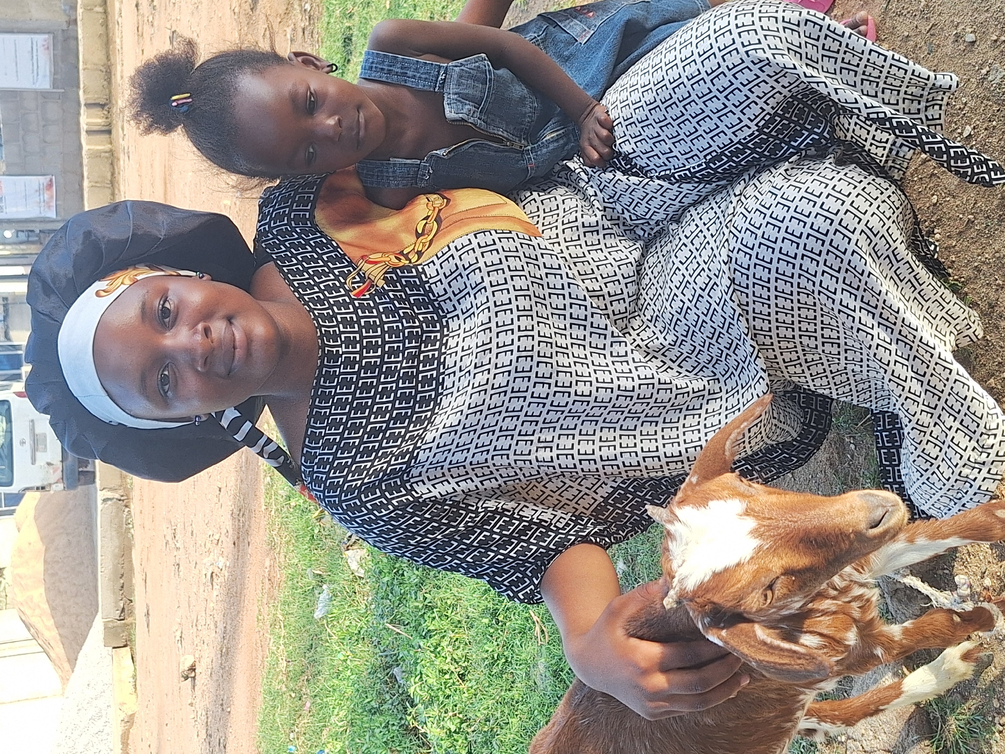 A woman wearing a black head covering and a black-and-white patterned garment, along with a young girl wearing denim overalls, are sitting together on the ground in an outdoor setting. The woman is holding a brown-and-white goat. The ground consists of both dirt and green grassy areas. A building and a white vehicle can be seen in the background, suggesting an urban environment within the city of Sabon Tasha, Nigeria.