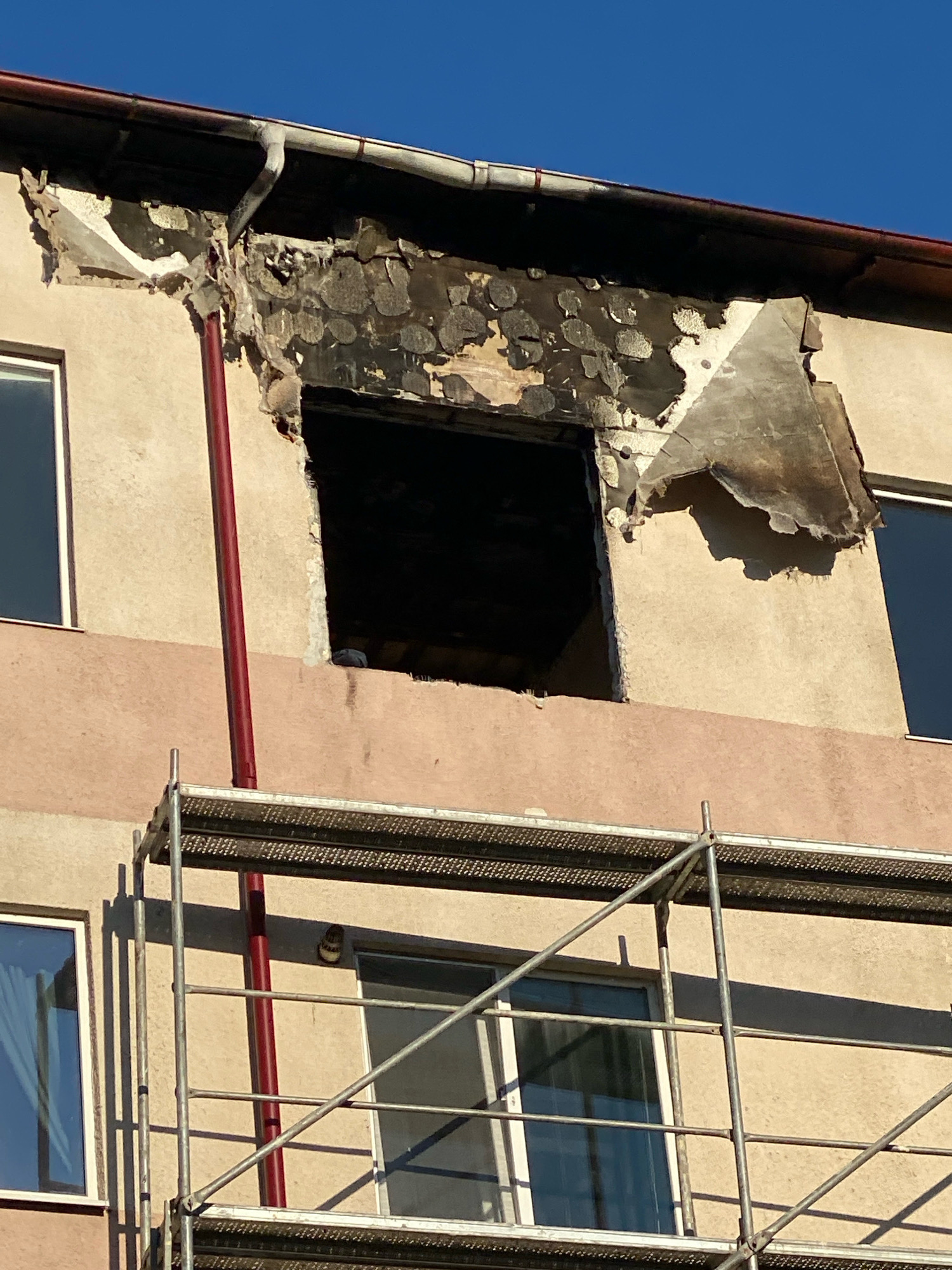 A close-up, low-angle shot shows damage to the facade of a building, likely an apartment block, under a clear blue sky. Scaffolding is erected in front of the building, obscuring the lower floors and obscuring some of the damaged area. The upper section of the building exhibits significant fire damage, with the facade blackened and peeling away, revealing charred material underneath. A large, rectangular opening, consistent with a damaged window or doorway, is visible in the burnt section. A dark red downspout runs vertically along the facade, passing through the damaged area. The building's exterior is painted in two tones: a lighter beige on the upper section and a salmon pink on the lower section. Windows with dark frames and reflective glass are visible on both floors. The scene is suggestive of recent fire damage and ongoing repair work.