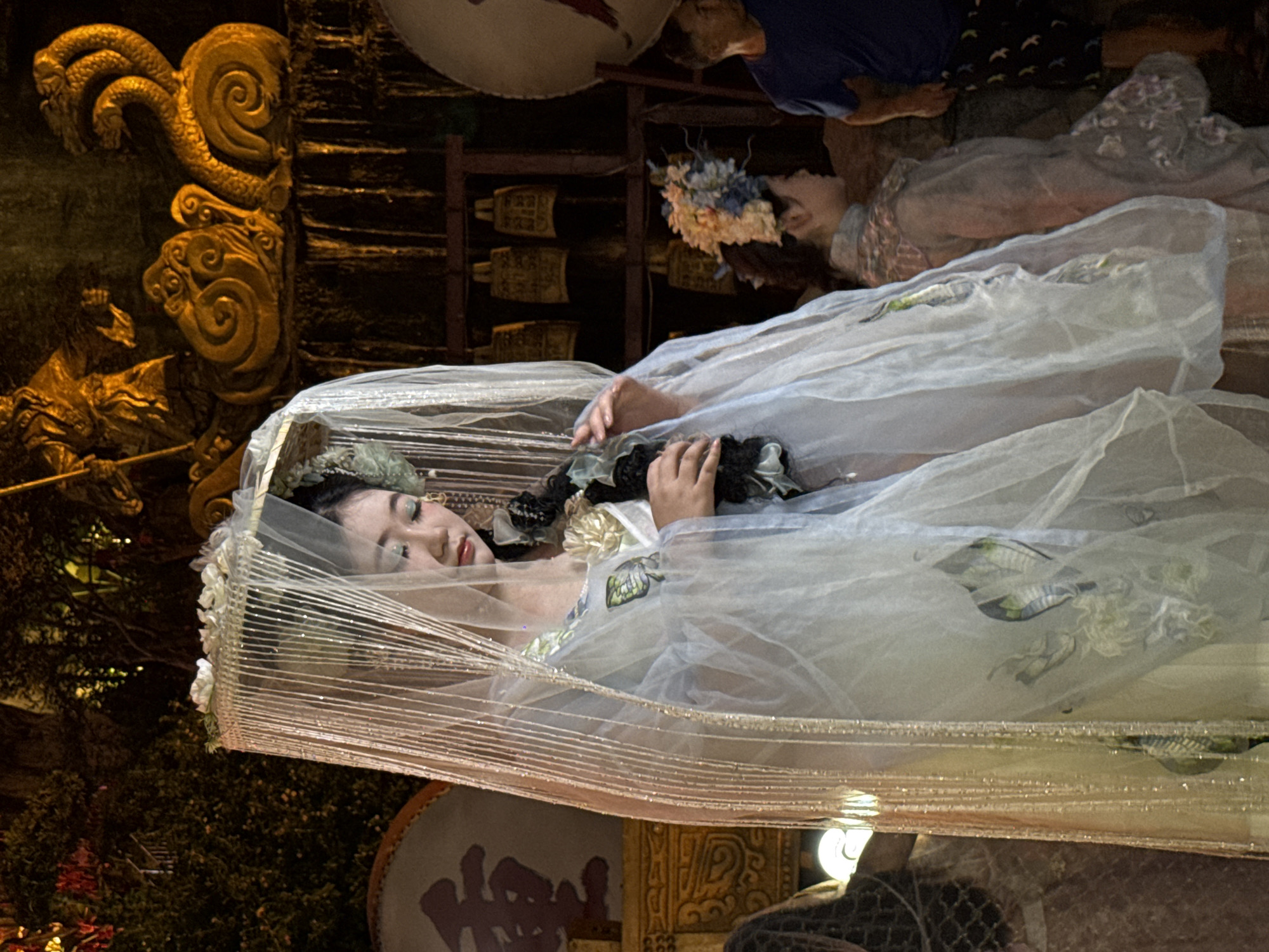 In Yuzhong District, China, a young Asian woman is the central focus of this dimly lit scene, possibly a performance or cultural event. She is adorned in a pale blue, sheer garment with floral and butterfly motifs, draped over a lighter undergarment. A delicate, beaded veil with floral embellishments cascades from her headpiece, partially obscuring her face and the elaborate braid of her dark hair. Her makeup, with blue eyeshadow and red lipstick, is prominent. She holds a dark object, possibly a prop, in her hands.
Behind her, a large, round drum with a red Chinese character is visible. Further back, a golden, sculpted mural depicts a multi-headed serpent coiled around a decorative cloud formation. Hanging bells are also visible in the background. To the right, another woman in a floral headdress and a translucent garment stands in profile. In the far right of the frame, a man in a blue t-shirt and patterned shorts is partially visible. The environment appears to be outdoors at night, with warm lighting illuminating the performers and decorations.