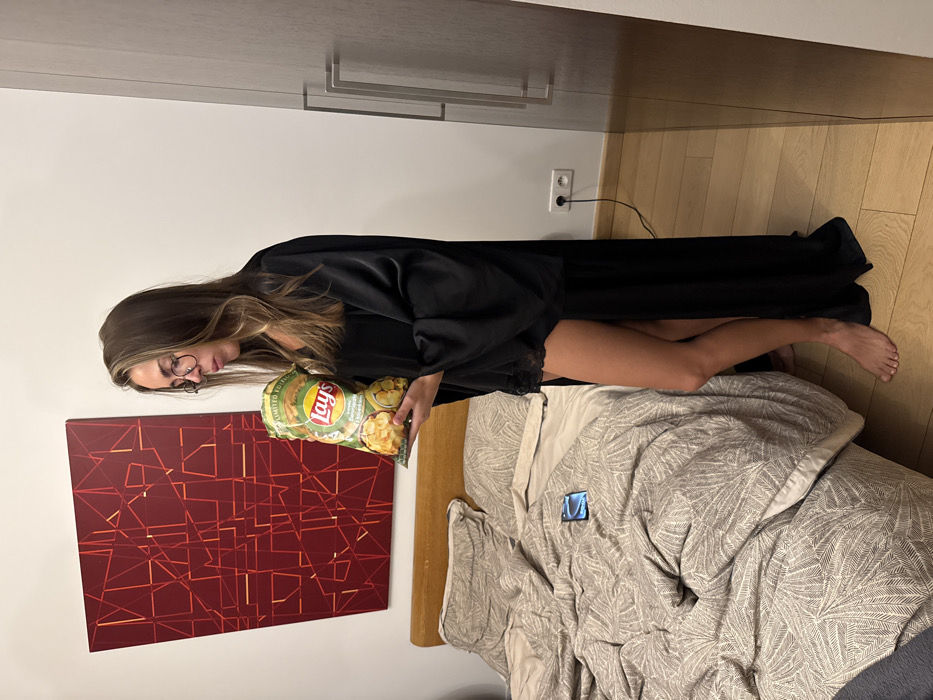 A woman stands in a bedroom next to a bed covered in a patterned gray comforter. She wears glasses and a long black robe, and holds a bag of Lay's potato chips. A red abstract painting hangs on the wall behind her. The room has light wood floors and built-in cabinets. An electrical outlet is visible on the wall. A blue object is also on the bed.