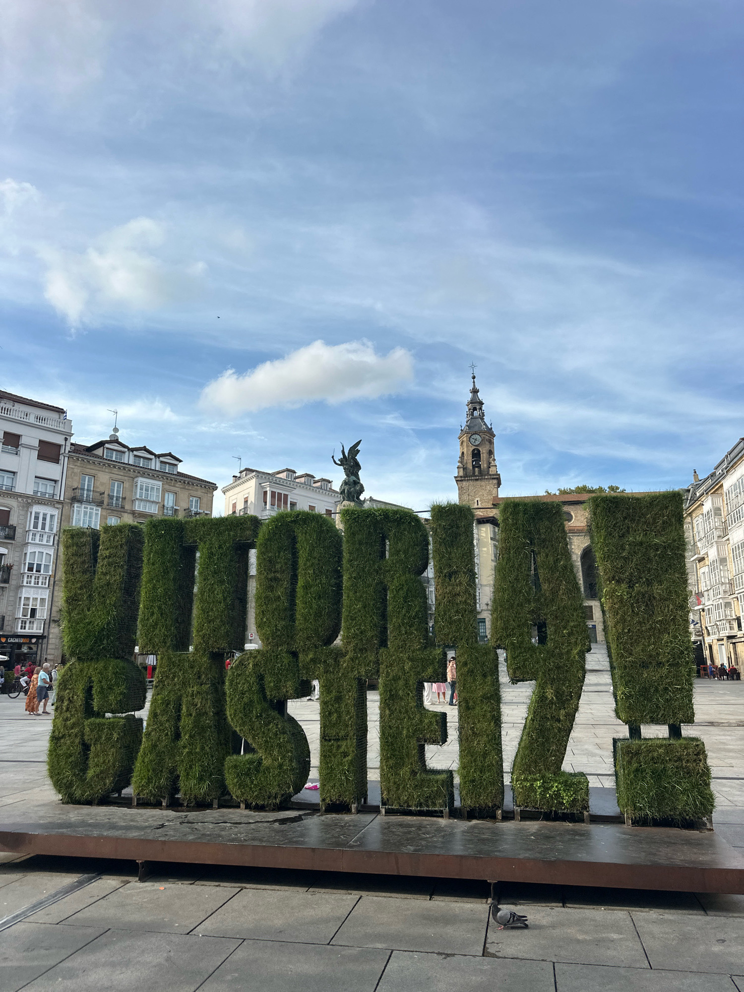 The image depicts a large, green, grass-covered sculpture spelling out "VITORIA-GASTEIZ!" in an outdoor public square. The sculpture is positioned in the foreground, occupying the majority of the frame. Behind the sculpture, a city square is visible, paved with grey stone tiles. Several buildings with a European architectural style, featuring balconies and multiple stories, line the square. In the background, a prominent clock tower stands tall, with a church spire visible above it. The sky is partly cloudy with a blue hue. A few individuals are scattered throughout the square, some walking and others appearing to be seated. A pigeon is also present in the foreground, standing on the tiled ground. The overall scene suggests a daytime setting in a European city.