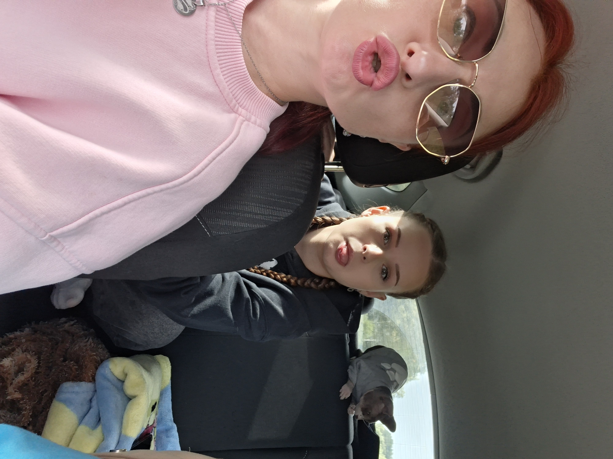 In a vehicle, a woman with red hair and sunglasses is leaning forward, puckering her lips and making a kissing face. Beside her, a young woman with braided hair is sticking out her tongue, looking at the camera. Behind them, a small chihuahua dog is wearing a grey sweater and lying on the back seat. The interior of the car is visible, with seats and a headrest. The background outside the car shows greenery and trees, suggesting a rural setting, possibly on the outskirts of Sloboda Rural Council, Belarus, during daylight hours.