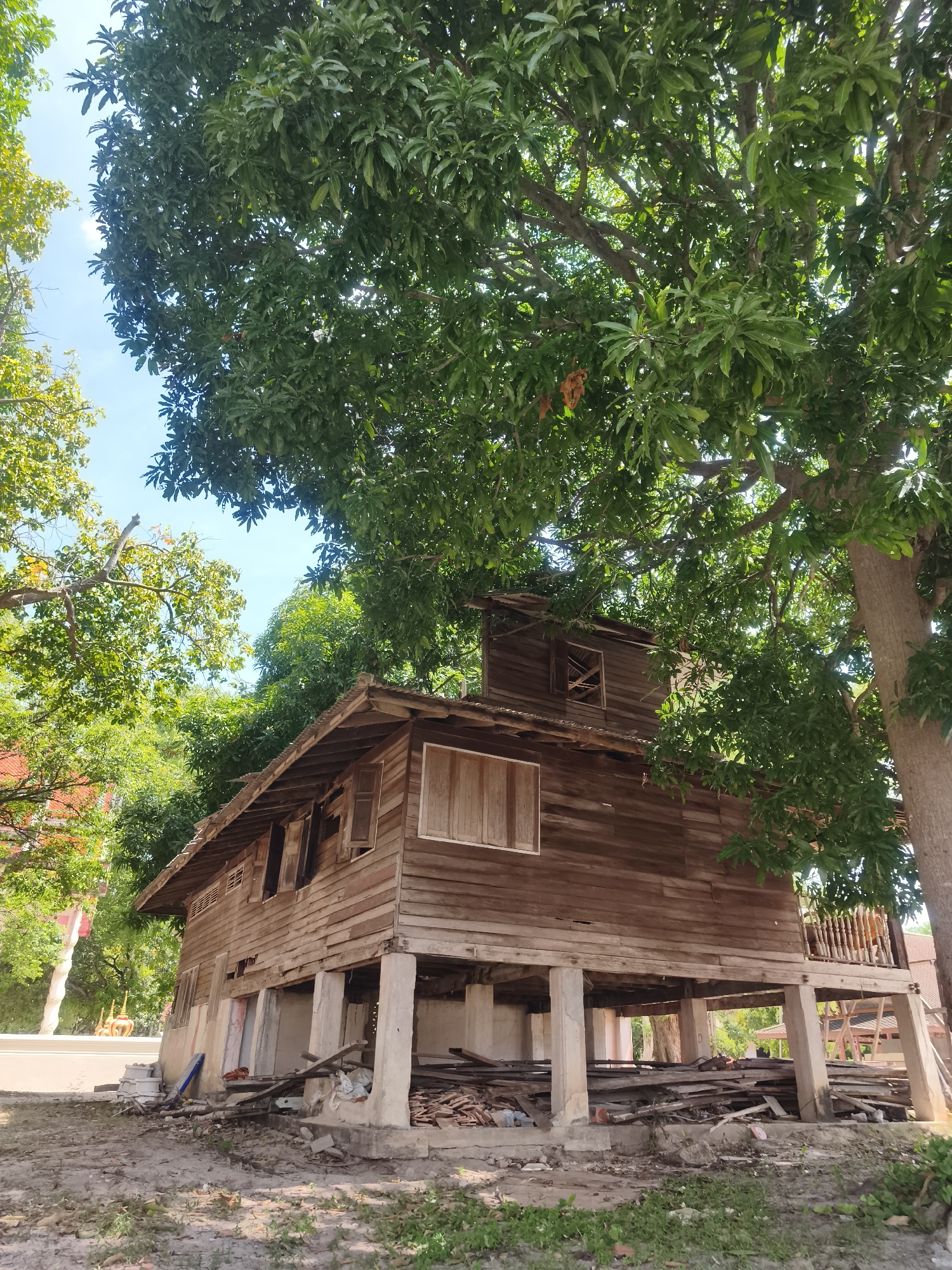The image depicts an old, weathered wooden stilt house located outdoors, under the shade of a large tree with dense green foliage. The house appears to be in a state of disrepair, with visible damage to its wooden structure and a dilapidated roof. The stilt foundation supports the elevated structure, and the area beneath the house is cluttered with debris, including scattered wooden planks, bricks, and various discarded materials. The ground around the house is uneven and appears to be a dirt or unpaved area with sparse vegetation. The sky is visible through the tree canopy, suggesting an outdoor setting. There are no people visible in the image. The context provided suggests this location is in the Ko Samui District of Thailand.