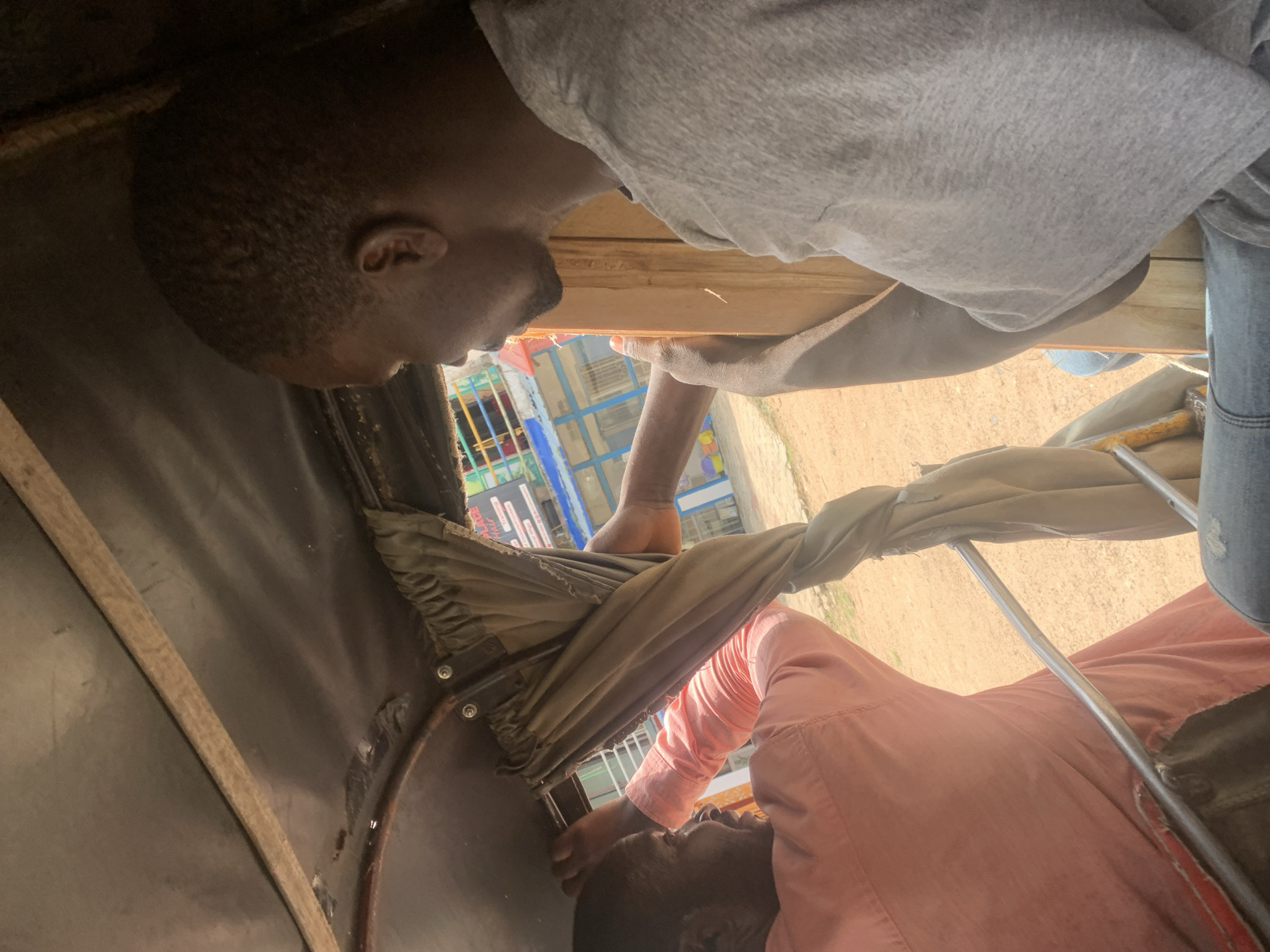The image shows two men inside what appears to be the passenger area of a motorized tricycle, commonly known as a "Keke Napep" in Nigeria. The perspective is from inside the tricycle, looking upwards.
The first man, on the left, is wearing a grey t-shirt and has short hair. He is leaning back, his head is partially visible. His face is turned away from the camera.
The second man, on the right, is wearing a light pink collared shirt. He is leaning forward and looking downwards. His face is visible, and he has a dark complexion.
The interior of the tricycle features dark fabric lining and some metal framing. A beige fabric, possibly part of a cover or canopy, is draped across the middle of the frame, obscuring some of the view. The exterior of the tricycle is visible through openings, showing a dusty, outdoor environment. In the background, there are glimpses of buildings and possibly market stalls with colorful elements, suggesting an urban or semi-urban setting.
The lighting suggests it is daytime, likely with bright sunlight given the outdoor setting and shadows. The overall scene conveys a sense of everyday travel or transport within a Nigerian context. No visible text is discernible.