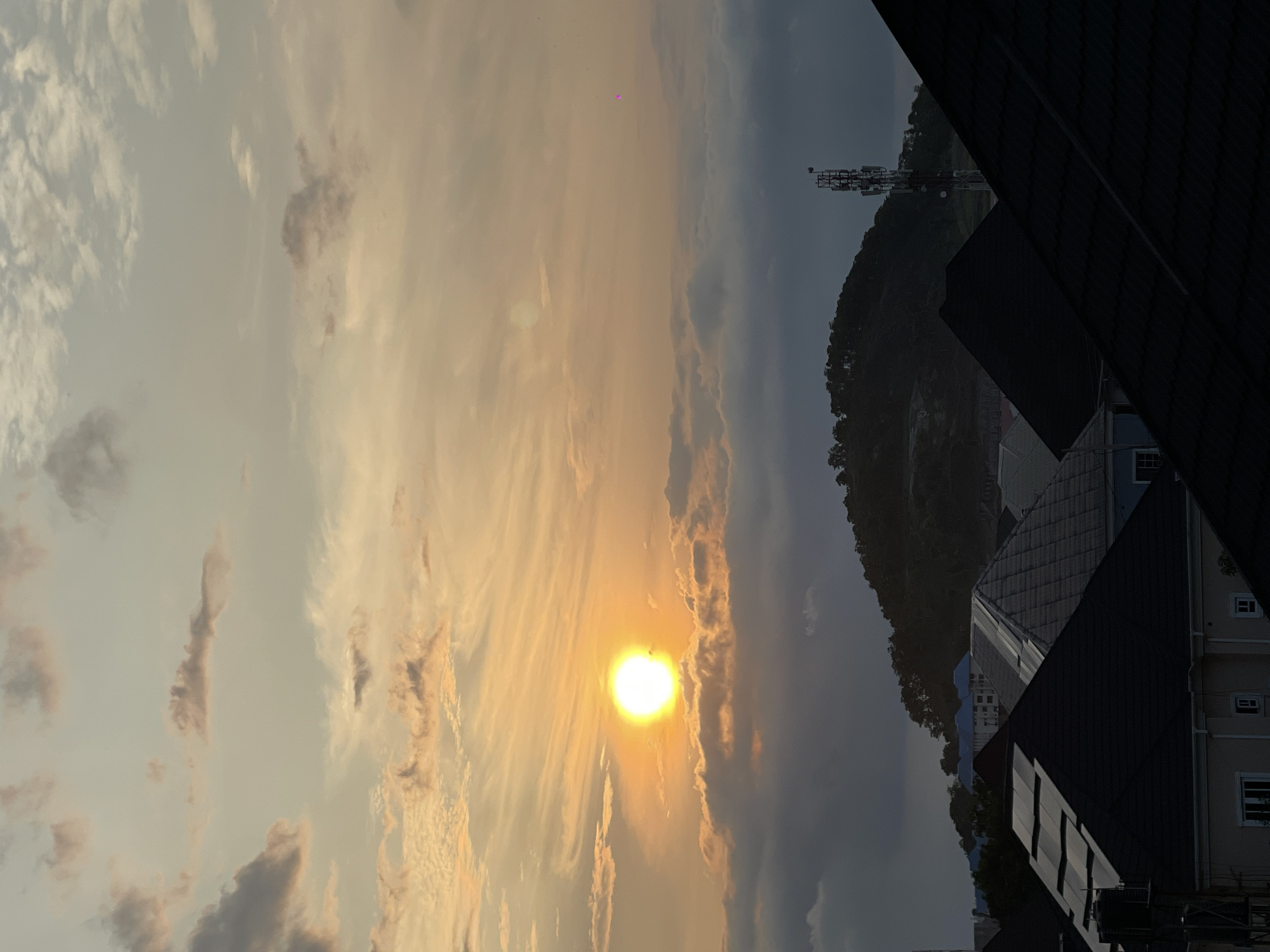 The image depicts a sunset over a residential area in Abuja, Nigeria. The sun, a bright orange orb, is partially obscured by clouds, casting a warm glow across the sky, which is a mix of soft yellows, oranges, and muted blues, with wispy clouds scattered throughout. In the mid-ground, a densely forested hill rises, topped with trees. A cellular tower stands prominently on the right side of the hill. In the foreground, the dark, angular roofs of houses are visible, with some structures showing blue-tiled roofs. The overall impression is a serene, urban landscape transitioning into evening.