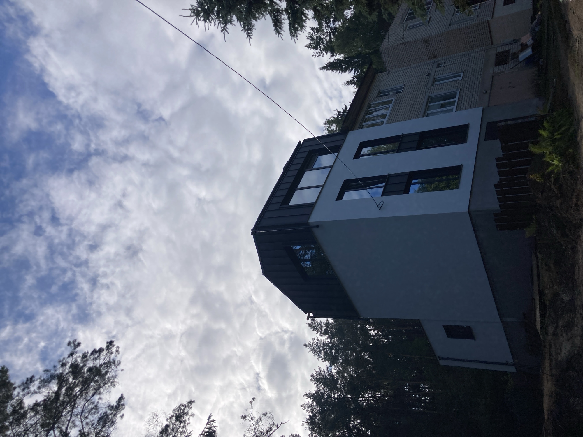The image depicts a modern, multi-story building with a dark, sloped roof and large, reflective windows. The building appears to be under construction or recently completed, with unfinished elements visible at the base. It is situated in a wooded area, with tall coniferous trees surrounding it. In the background, another, older brick building is visible. The sky is cloudy, with patches of blue visible. The scene is set in Astrashitskagaradokski rural council, Belarus. There are no people or animals visible in the image.