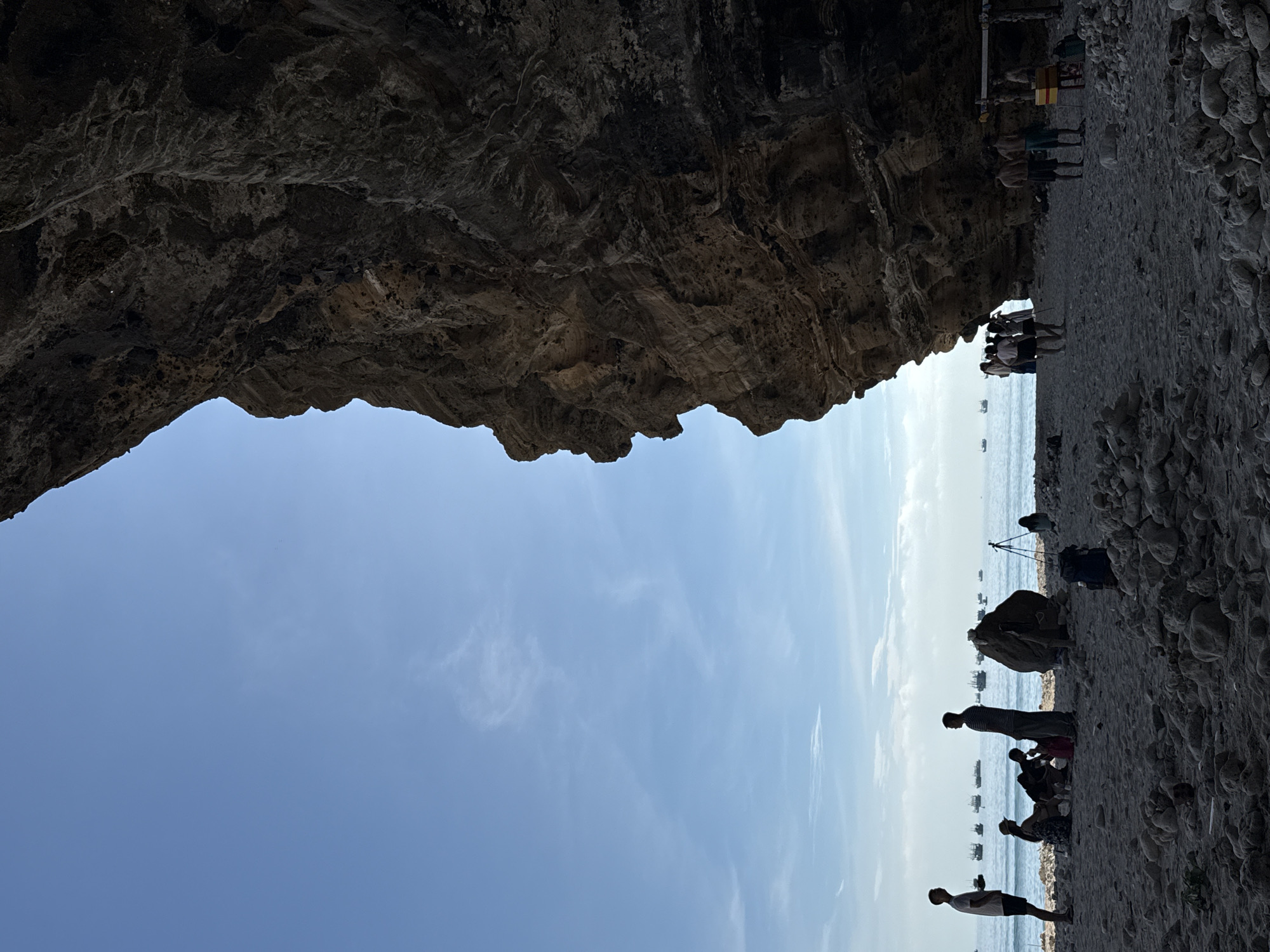 A wide shot captures a beach scene in the Lý Sơn Special Administrative Region, Vietnam. The foreground is dominated by a large, textured rock formation casting a shadow over the sand and a group of people. The beach is covered in pebbles and sand, with the ocean visible in the background. A clear blue sky with wispy clouds stretches above, and numerous boats are dotted across the water. Several people are scattered across the beach; some are standing, others are sitting, and a small group appears to be interacting near a makeshift shelter. One individual stands near the water's edge, while others are gathered in smaller clusters on the sand. A tripod and a backpack are visible on the beach, suggesting recreational activity. The overall atmosphere is one of a relaxed coastal outing.