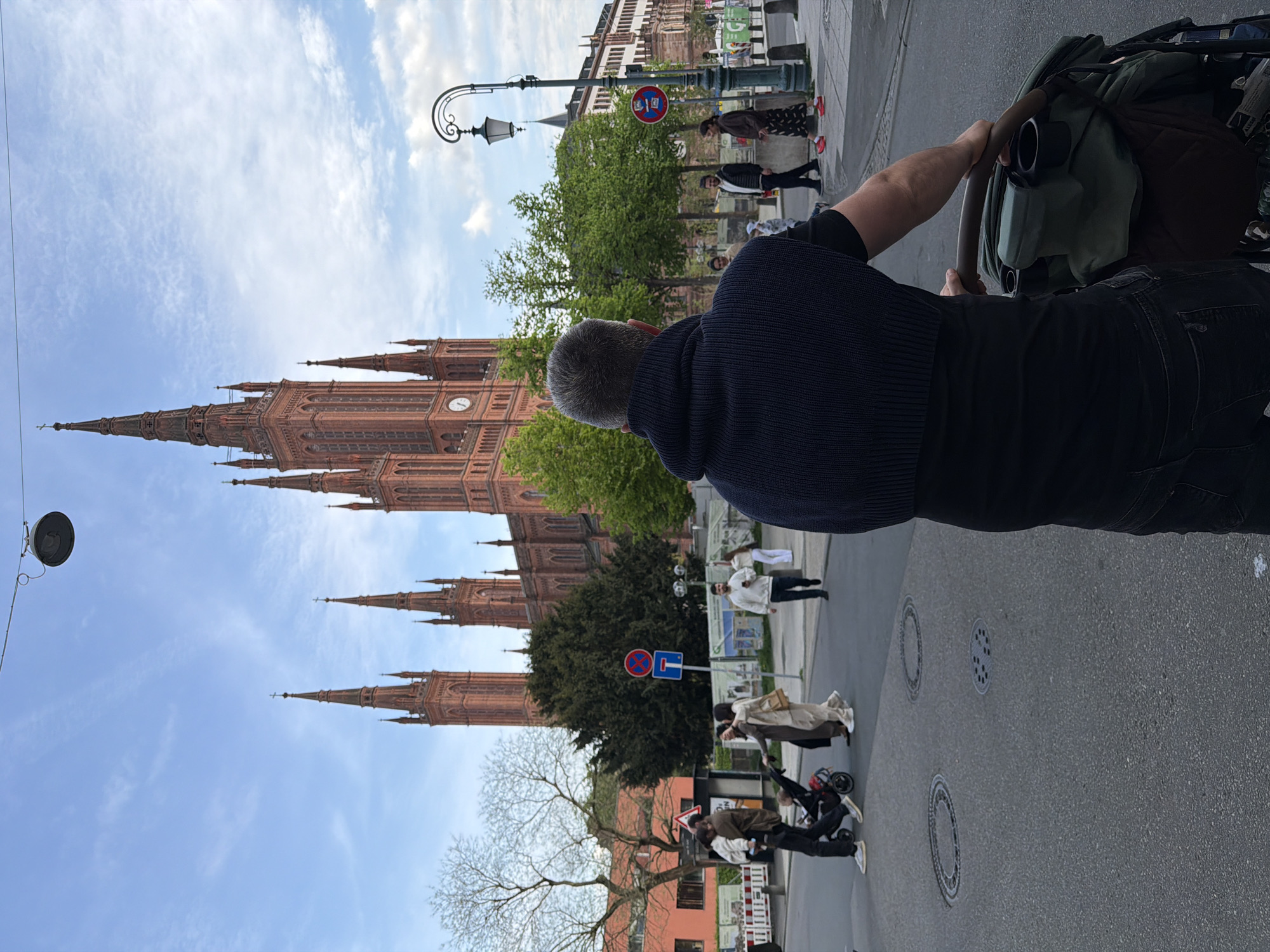 A man pushing a stroller walks along a paved street in Wiesbaden, Germany. The Market Church (Marktkirche) is visible to the left, its red brick facade and spires reaching towards a blue sky with scattered clouds. Trees line the street, and several pedestrians can be seen walking in the background. Streetlights and traffic signs are visible along the side of the road. The man is wearing dark clothes, and the stroller has a green bag attached. The camera's perspective is from behind the man, close to the stroller.