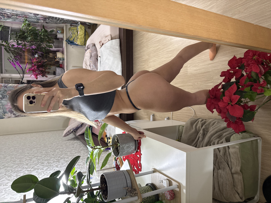 A slender woman poses for a mirror selfie, wearing a gray top and thong. Her phone and smartwatch are visible. The background shows a bedroom with a bed, plants, and storage. Potted plants surround the bottom of the mirror, while a red poinsettia stands near the edge on the right. A white cabinet is visible next to the poinsettia. The room is brightly lit.