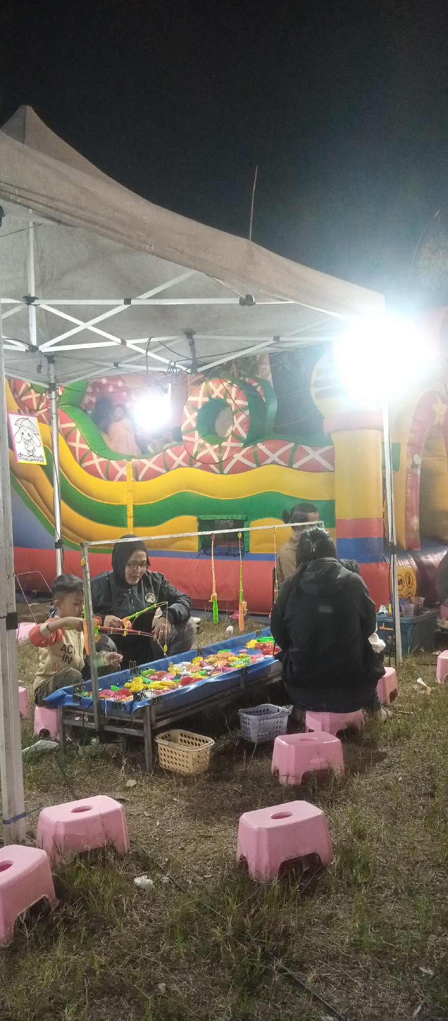 The image depicts an outdoor game stall at night, possibly at a festival or fair, in Cermo, Indonesia. A large, inflatable structure with colorful wavy patterns, primarily yellow, green, and red, is visible in the background. Underneath a white tent, a group of people are gathered around a long, shallow blue trough filled with brightly colored plastic toys, resembling a fishing game.
In the foreground, a young boy in a yellow and red shirt is actively engaged in the game, holding a fishing rod. Beside him, an adult wearing a dark hooded jacket and glasses is also participating. To the right, another adult is seated with their back to the camera, also facing the game. Several pink plastic stools are scattered on the grassy ground around the game area, with two baskets placed near the trough. The lighting suggests artificial illumination from the tent and possibly the inflatable structure, creating a warm glow against the dark night sky. The ground is covered in dry grass and some litter.