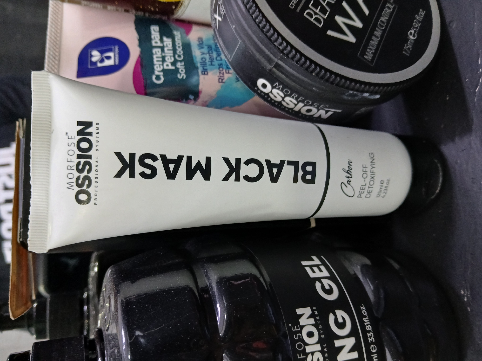 The image shows a collection of personal care products. Dominating the foreground is a white tube labeled "MORFOSE OSSION PROFESSIONAL SYSTEMS BLACK MASK," further described as "Carbon PEEL-OFF DETOXIFYING," with a volume of 125ml or 4.23fl.oz. Partially visible behind it are other products, including a pink and blue tube labeled "Crema para Peinar Soft Coconut" and a black circular container labeled "BEA W," with "MAXIMUM CONTROL" and a volume of 175ml or 5.92 fl.oz. A partially visible black bottle is in the lower part of the frame labeled "MORFOSE SION ONAL SYSTEMS NG GEL" with a volume of "mle 33.81fl.oz." The scene appears to be a still life of these products placed close together, with a dark background or surface underneath. There is no indication of a location in Juan Griego, Venezuela, or any people present.