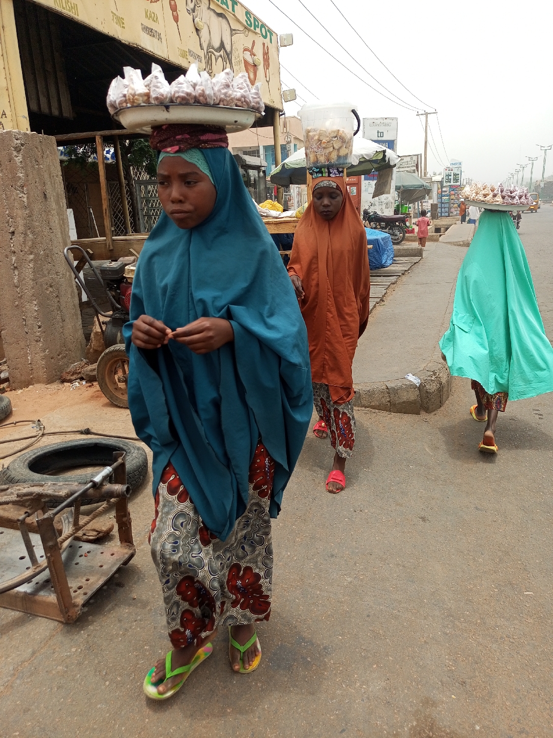 Three women are walking on a street in Gombe, Nigeria. Each woman carries goods on her head. The woman in the foreground wears a blue hijab and a patterned skirt, and carries a tray of wrapped items. The woman behind her wears an orange hijab and carries a container of food. The woman in the background wears a green hijab and carries a tray of items. The street is paved and there are buildings and vehicles visible in the background. There is a metal structure and a tire on the side of the street in the foreground.