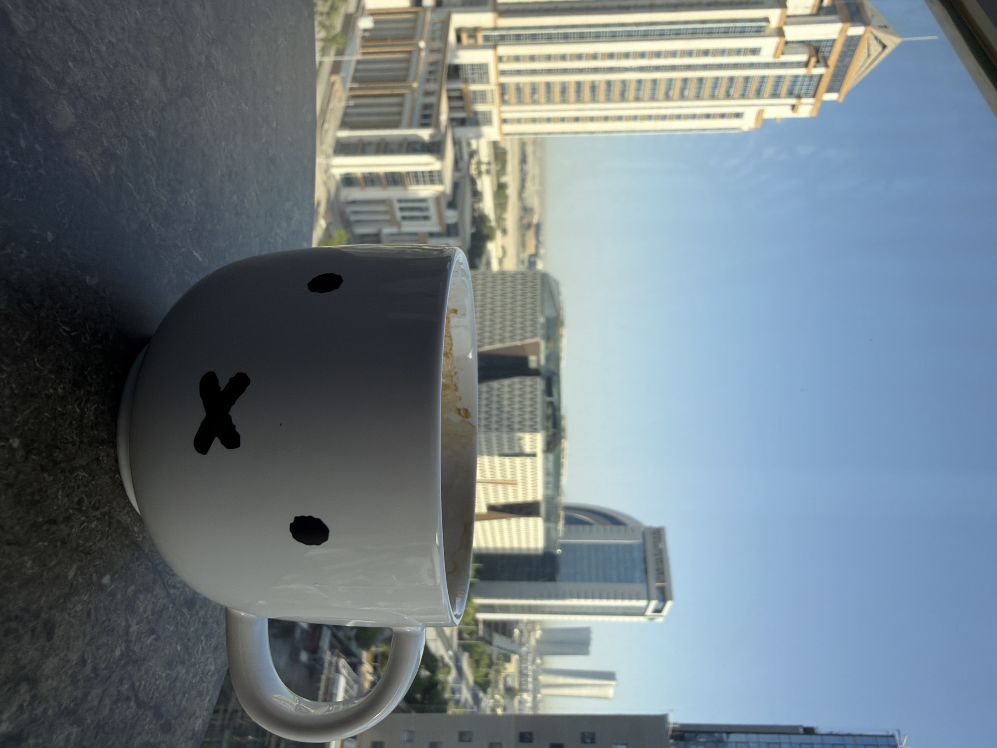 A white ceramic mug with a Miffy-like face, featuring a black dot for an eye and an "X" for a mouth, sits on a dark textured surface. The mug contains a light brown liquid, likely coffee, with some residue on the rim. In the background, seen through a window, is a cityscape with several modern buildings under a clear blue sky. One prominent tall, light-colored building with gold accents stands to the left, and other buildings of varying heights and architectural styles are visible in the distance. The scene appears to be a view from a high-rise apartment or office, suggesting an urban environment in Unknown, Unknown. The lighting suggests it is daytime, likely morning or afternoon.