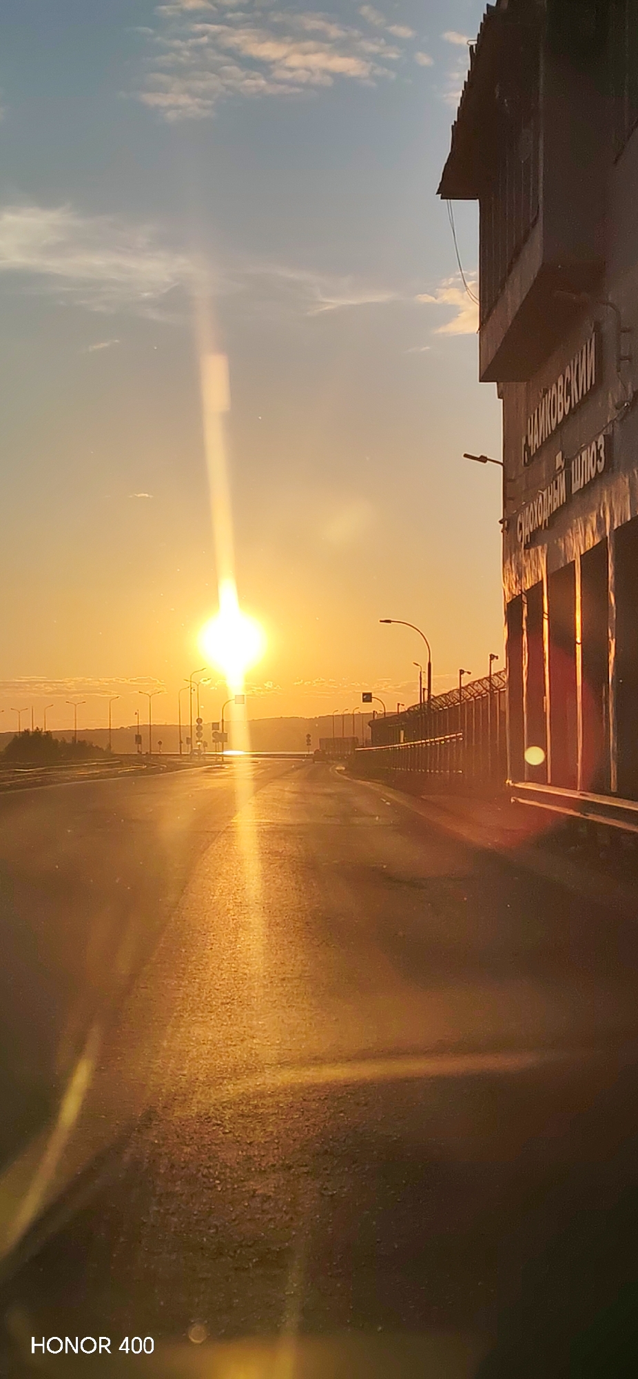 An outdoor, vertically oriented photograph captures a low-angle sun over an asphalt road, facing towards the horizon, likely during sunrise or sunset. The intense, bright yellow sun, positioned in the middle left, casts a strong vertical lens flare that extends across the sky and reflects prominently down the wet-appearing road surface.
To the immediate right, a large, dark, utilitarian building with a segmented facade and an overhanging roofline runs parallel to the road. White Cyrillic lettering on the building clearly spells out "ЧАЙКОВСКИЙ" (Chaikovsky) and "СУДОХОДНЫЙ ШЛЮЗ" (Navigation Lock), identifying the location as the Chaikovsky Navigation Lock in the city of Chaikovsky, Russia. The building's surface shows reflections of the low sun.
The road, possibly a bridge or elevated section, is devoid of vehicles or pedestrians. A series of streetlights with curved poles line both sides of the road in the mid-distance. Along the right side of the road, between the asphalt and the lock building, is a fence-like structure topped with barbed wire. Hilly terrain or elevated land is visible on the distant horizon under a hazy sky, which transitions from bright orange and yellow near the sun to a pale blue with scattered clouds higher up. The bottom left corner of the image contains a "HONOR 400" watermark.