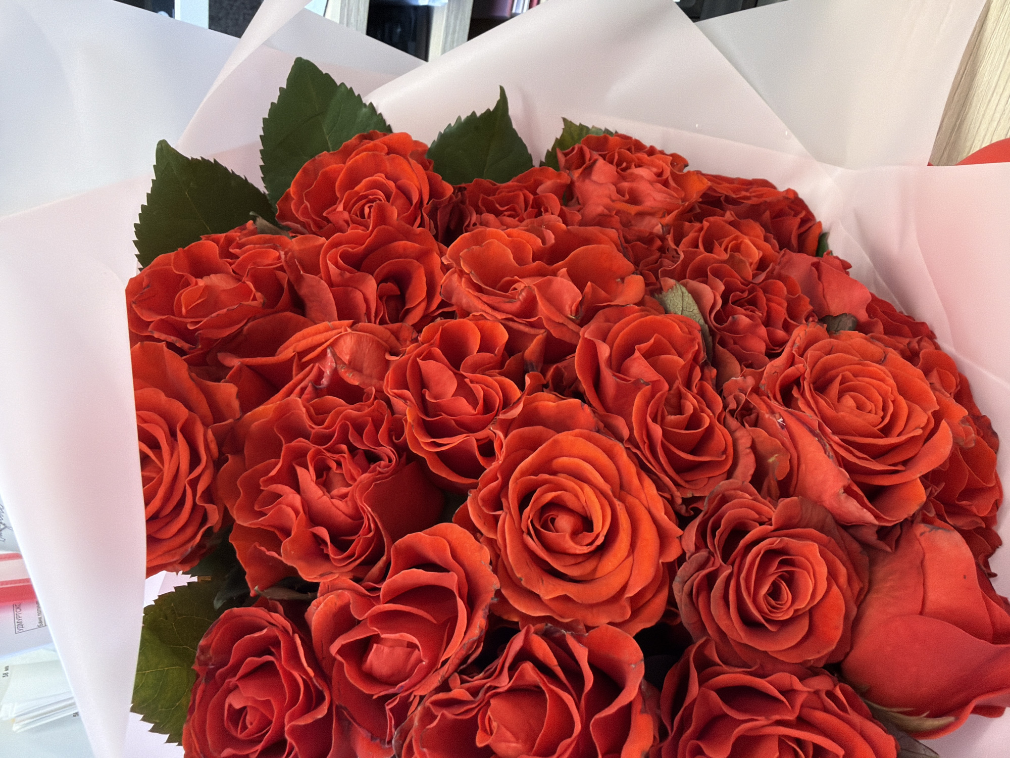 A bouquet of red roses is displayed wrapped in white paper. There is a desk or table to the side of the bouquet, which is partially obscured by the paper. The precise location in Izhevsk, Russia is not discernible from the image.