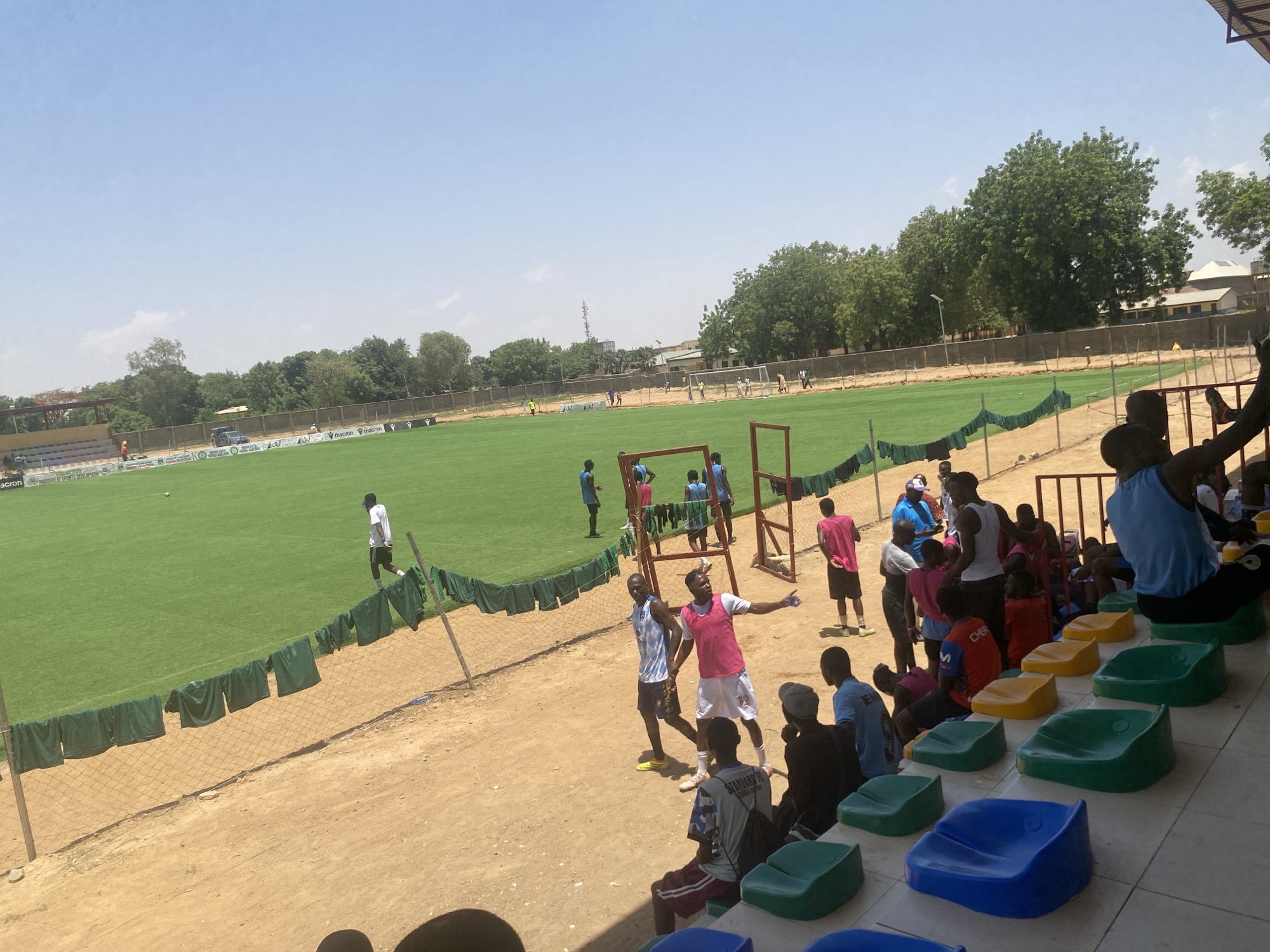 The image depicts a daytime scene at a soccer stadium in Tudun Wada, Nigeria. A green artificial turf field is visible, surrounded by a fence with green cloths draped along it. Several soccer players are on the field, some in white shirts and others in blue. Beside the field, on a dirt ground area, a small group of players and spectators are standing and sitting on multicolored stadium seats. One man is wearing a pink vest over a white shirt, gesturing with his right hand, while another nearby is in a light blue and white jersey. The sky is clear and bright, with trees and some buildings visible in the background, suggesting an urban environment.