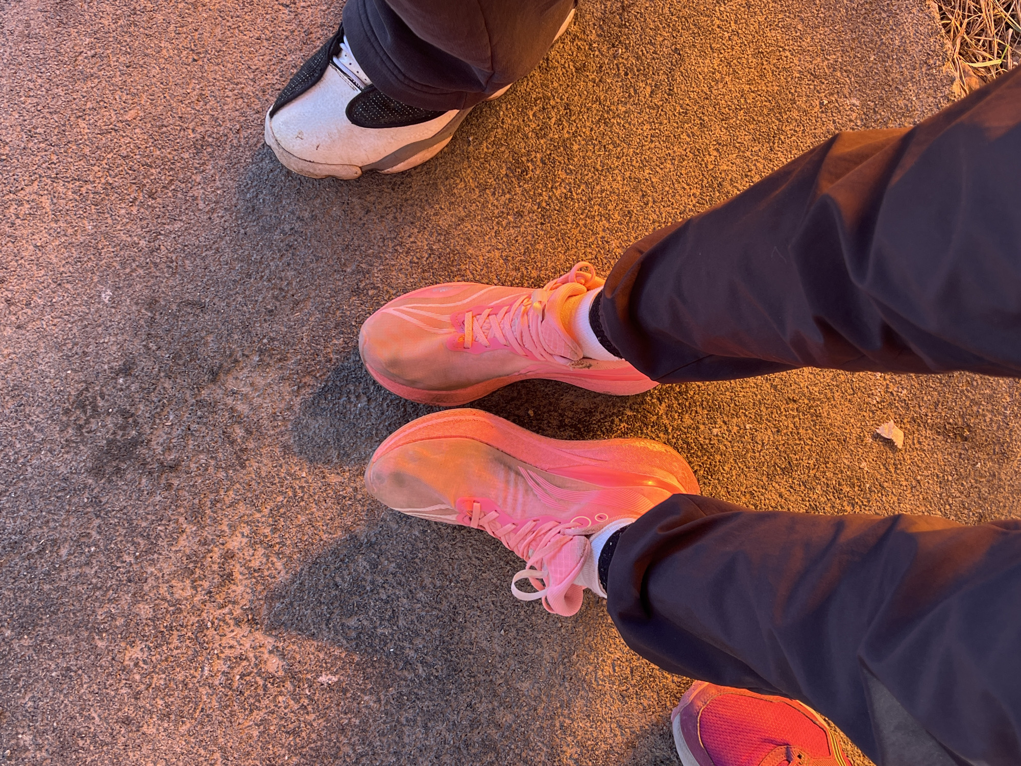 The image shows a street surface with three pairs of shoes visible. One pair is white and gray sneakers, worn by a person whose legs are partially visible at the top of the image. Another person stands wearing pink running shoes and dark pants. A small portion of the person's other shoe is visible in the bottom right of the frame. The street surface is textured. The lighting suggests it might be twilight or artificial lighting. Gory, Russia, is not discernible from the image.