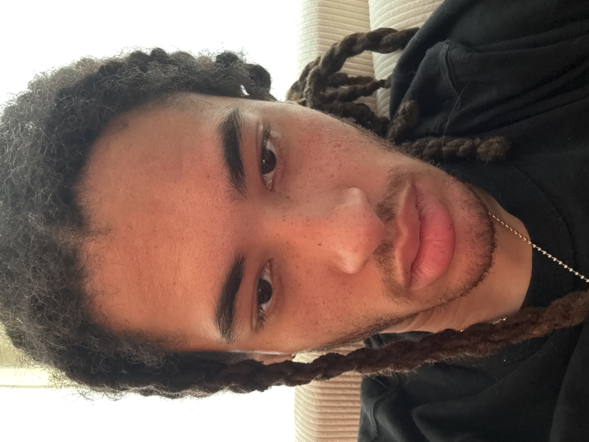 A close-up, slightly angled headshot of a young, fair-skinned person with dreadlocks. The hair is dark and appears to be partially styled into twists. The person has dark eyebrows, eyes, and a short, patchy beard. The person is wearing a dark-colored garment, and a silver chain necklace is visible. The background is blurry and beige in color. No information suggests this image was taken in Paris, France.