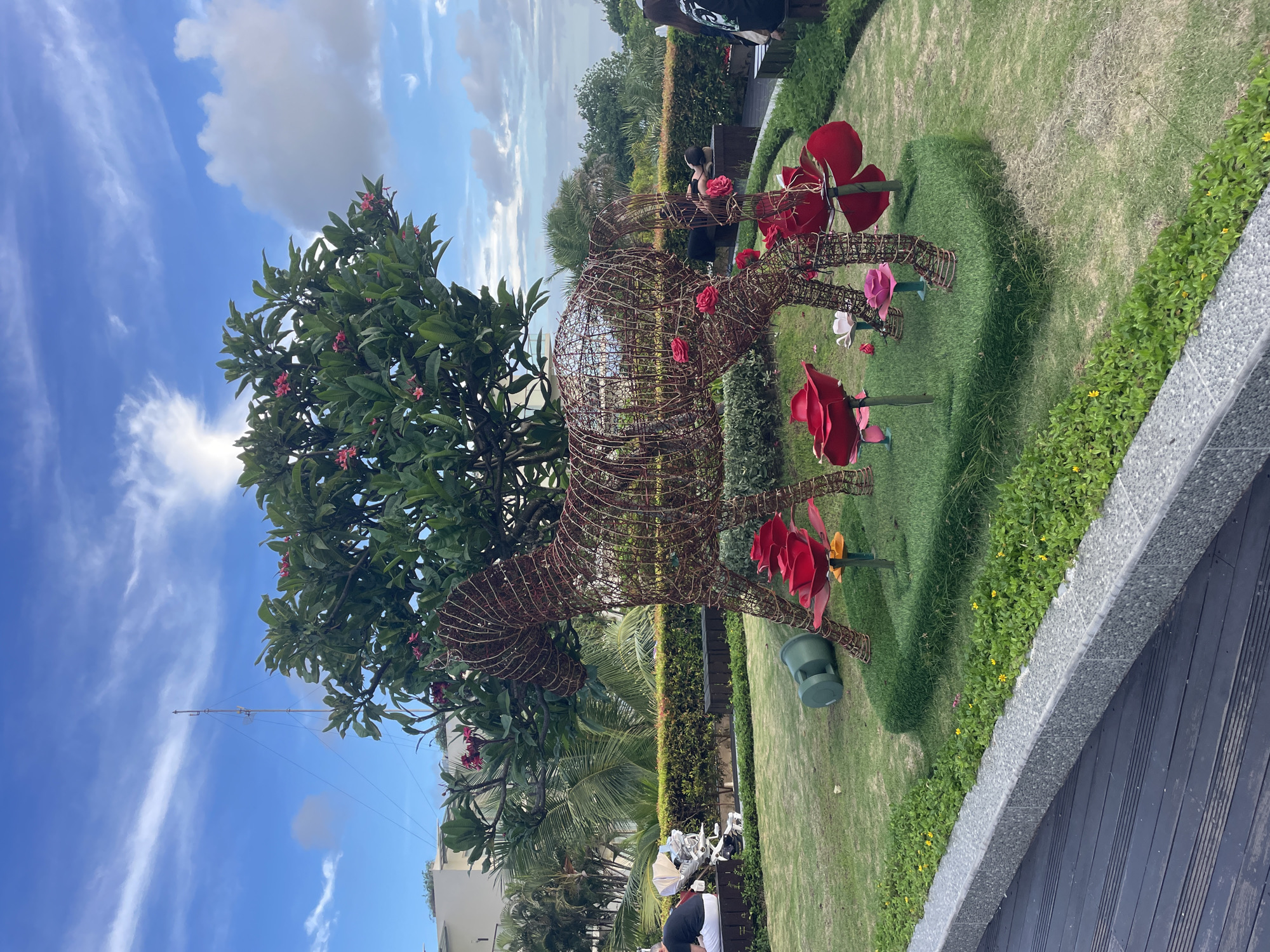 The image captures an outdoor garden or park setting on a bright, sunny day. A large, intricate wire-frame sculpture of an animal, possibly a deer or an abstract creature, is centrally located on a well-maintained grassy area. The sculpture is adorned with several artificial red and pink flowers, and additional large, artificial red and pink flowers are staked into the grass around it. A large tree with lush green leaves and small pink blossoms stands prominently on the left side of the frame, reaching towards the clear blue sky, which features scattered white clouds.
In the foreground on the right, there is a gray textured wall or divider bordering a dark wooden deck or walkway. Beyond this, a person is partially visible, bent over. In the mid-ground, another person sits at a table with their back to the camera, near the wire sculpture. Further in the background, green hedges and various trees, including palm trees, create a verdant backdrop. A building and a tall antenna or mast are visible in the distance under the blue sky. A green outdoor speaker is also visible near the base of the wire sculpture. The scene suggests a pleasant and well-maintained leisure area, possibly part of a resort or public garden in a tropical or subtropical climate. No visible text is present in the image.