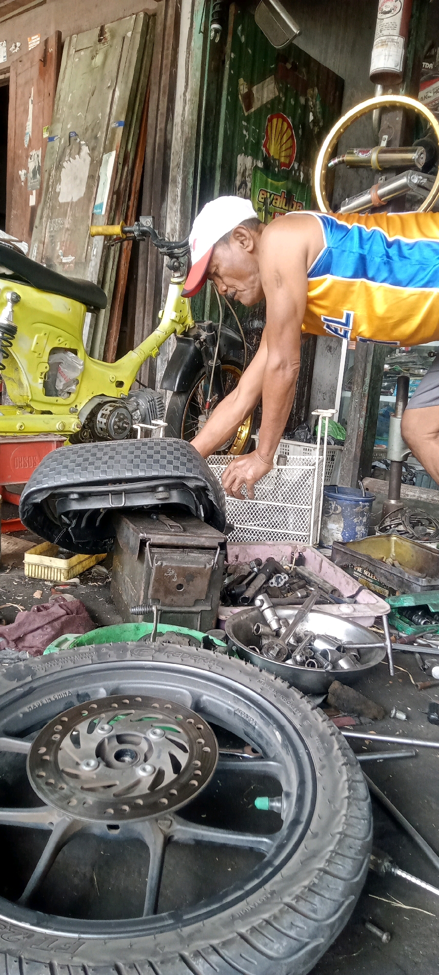 This is a medium shot of a mechanic working on a motorcycle in what appears to be an open-air workshop in Keteguhan, Indonesia. A middle-aged man wearing a white cap and a yellow and blue tank top is bent over, seemingly inspecting or repairing the front end of a bright yellow motorcycle. Tools and parts are scattered around him, including a disassembled motorcycle wheel in the foreground and an ammo box serving as a makeshift stand for the motorcycle seat. The background shows corrugated metal walls and various items typically found in a workshop, such as spare parts, containers, and what looks like a tire rim with exhaust pipes attached. The overall scene suggests a busy, functional workspace.