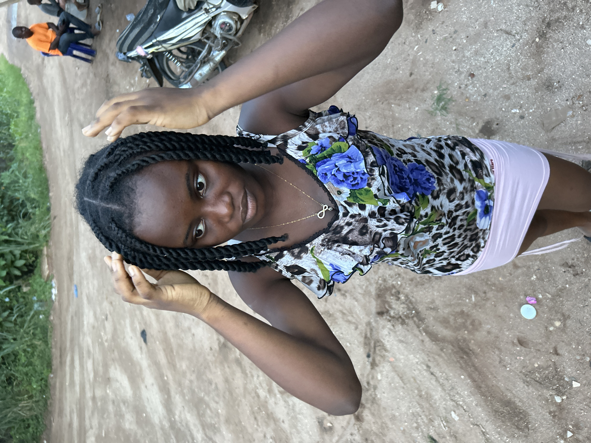 An eye-level shot captures a young woman posing outdoors on a dirt path in Ihiagwa, Nigeria. She is the central focus, looking directly at the camera with a neutral to slightly contemplative expression. Her hair is styled in dark braids, and she wears a sleeveless top featuring a leopard print pattern with prominent blue floral designs and ruffled trim. A delicate gold-colored necklace with an infinity symbol charm adorns her neck. Below the top, she wears a light pink mini-skirt, on the side of which a small white label with red text, partially legible as "STAR," is visible. Her hands are raised, resting near her braided hair.
The setting is an unpaved, dusty ground, indicative of a dirt road or path. On the left side of the frame, some green foliage or grass is visible. In the background, to the right, a motorcycle or scooter is parked. Further back on the left, two other individuals are present; one man is clearly visible, seated on a blue stool or chair, wearing an orange shirt and dark pants, while another person is partially obscured behind him. The lighting suggests it is daytime, under clear skies, with soft illumination. The overall scene depicts a casual moment in a semi-urban or rural outdoor environment.