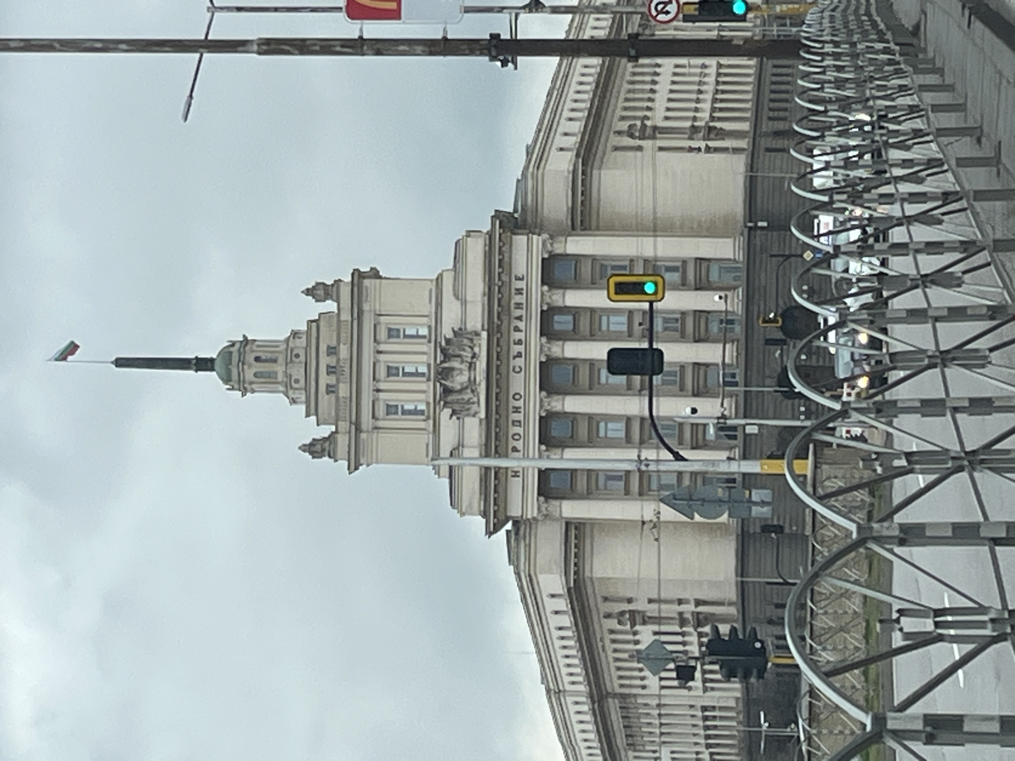 The image depicts the National Assembly of Bulgaria (Народно събрание) building in Sofia, observed from street level under an overcast sky. The large, light-colored building, located prominently in the mid-to-background, features classical architectural elements including columns, multiple windows, and decorative cornices. A central, multi-tiered tower culminates in a tall spire, from which the Bulgarian national flag, a white, green, and red tricolor, is flying. The Cyrillic text "НАРОДНО СЪБРАНИЕ" is visible on the building's facade.
In the midground, a multi-lane asphalt road with painted lane markings is visible, separated from the viewer's position by a metal pedestrian barrier. Several vehicles, primarily light-colored cars, are present on the road, suggesting active traffic flow. Multiple traffic lights are visible; one pole positioned in front of the building displays a green light, and another pole to the far right shows a green right-turn arrow. A tall utility pole on the right side of the frame supports a street light and a red and yellow McDonald's sign. Various street signs are also mounted on poles.
The immediate foreground features a grey metal barrier or fence, composed of repeated arched and criss-cross patterns, extending horizontally across the lower portion of the image. No individuals are clearly identifiable within the scene. The lighting conditions indicate a cloudy daytime environment.