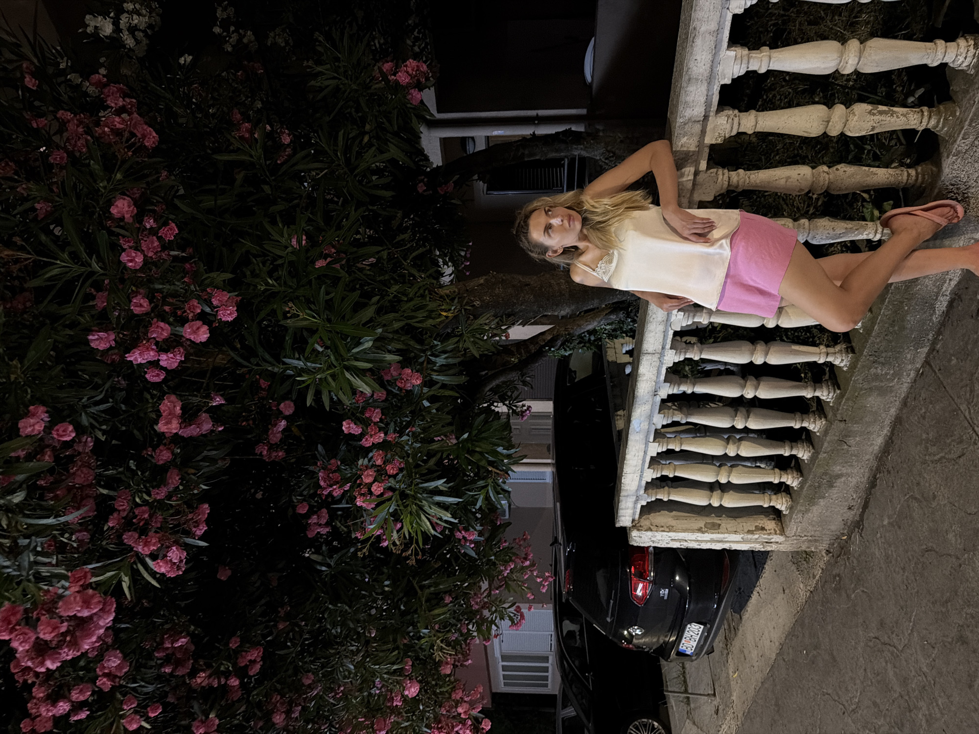 A blonde woman in a satin tank top and pink shorts leans on a stone balustrade at night in Budva, Montenegro. She is positioned under a blooming oleander tree with pink flowers, to the left of a black Volkswagen Golf parked on the street. The scene is illuminated by artificial light, casting shadows and highlighting the details of the woman, the car, and the floral display. A building with white-framed windows is visible behind the tree. The balustrade is aged and weathered, with ornate stone pillars. The license plate of the car reads "BD BH 203".