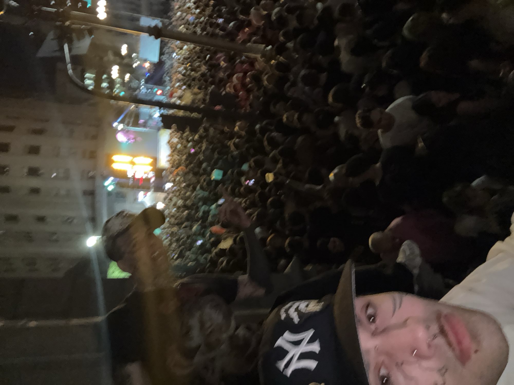 The image shows a large crowd of people at an event, likely a concert or festival, at night in Buenos Aires, Argentina. In the foreground, a person is taking a selfie, wearing a baseball cap with a "NY" logo. Other people are visible around the selfie taker, some with their arms raised. The background is filled with a dense crowd extending towards a stage with bright lights and video screens. Buildings are visible in the distance.