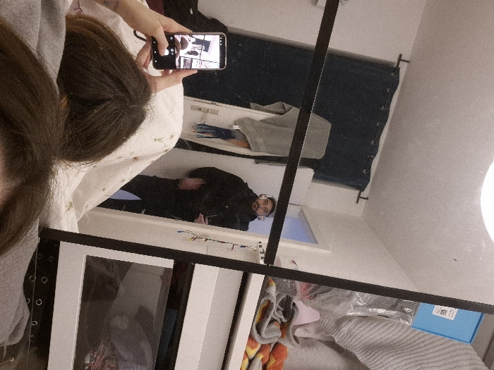 The image displays an indoor scene within a residence located in Strasbourg, France. The perspective is from a person lying in bed, showing the top of their head and their right hand holding a black smartphone. The smartphone's screen is active, displaying its camera interface which is capturing a live reflection of the room.
The primary subject of the reflection is a male figure, seen from the waist up, standing in a doorway. He wears a dark hooded garment and white over-ear headphones, with a visible beard. He appears to be looking towards the left within the reflection. To the left of the reflected male, a window with dark blue curtains is visible. The illuminated ceiling and a light fixture are reflected at the top of the image. To the right of the reflected male, a portion of white furniture or shelving holds a light-colored ribbed sweater, a blue box with a label, and other miscellaneous items. A string of colorful decorative lights is draped vertically along the right edge of the mirror frame or adjacent furniture. The person holding the phone is actively photographing this reflected scene. The bed is covered with a white duvet featuring a subtle green pattern and a grey blanket.