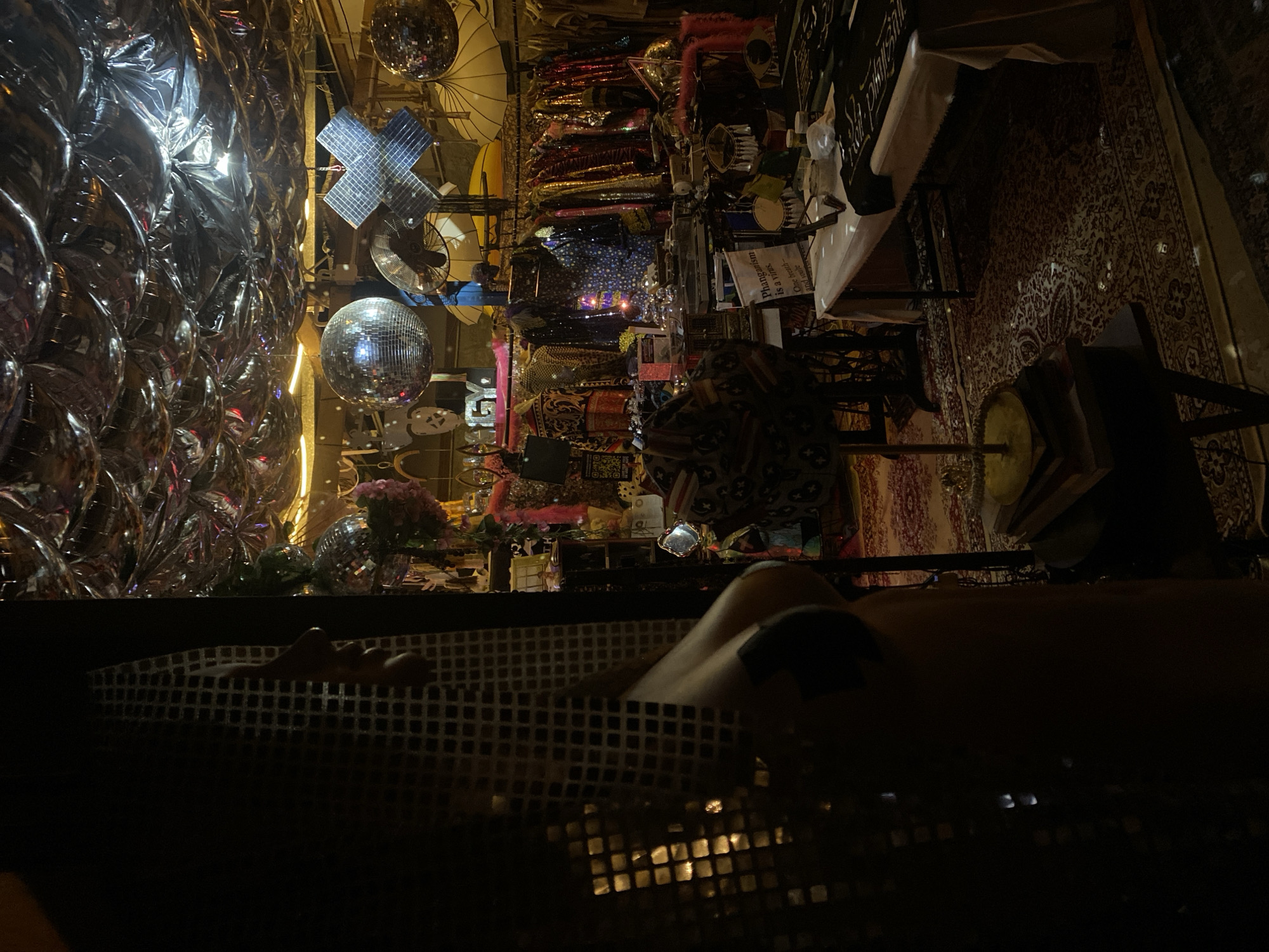 A low-angle, indoor shot shows a dimly lit space filled with decorations, objects, and a person in the foreground. On the left side of the image, a wall of metallic balloons reflects light. Further into the scene, disco balls hang from the ceiling, along with other decorative items. Clothing racks hold various garments. To the right, a carpet covers the floor. In the foreground, a person appears to be lying down. The location is indicated as being in Thong Sala, Thailand.
