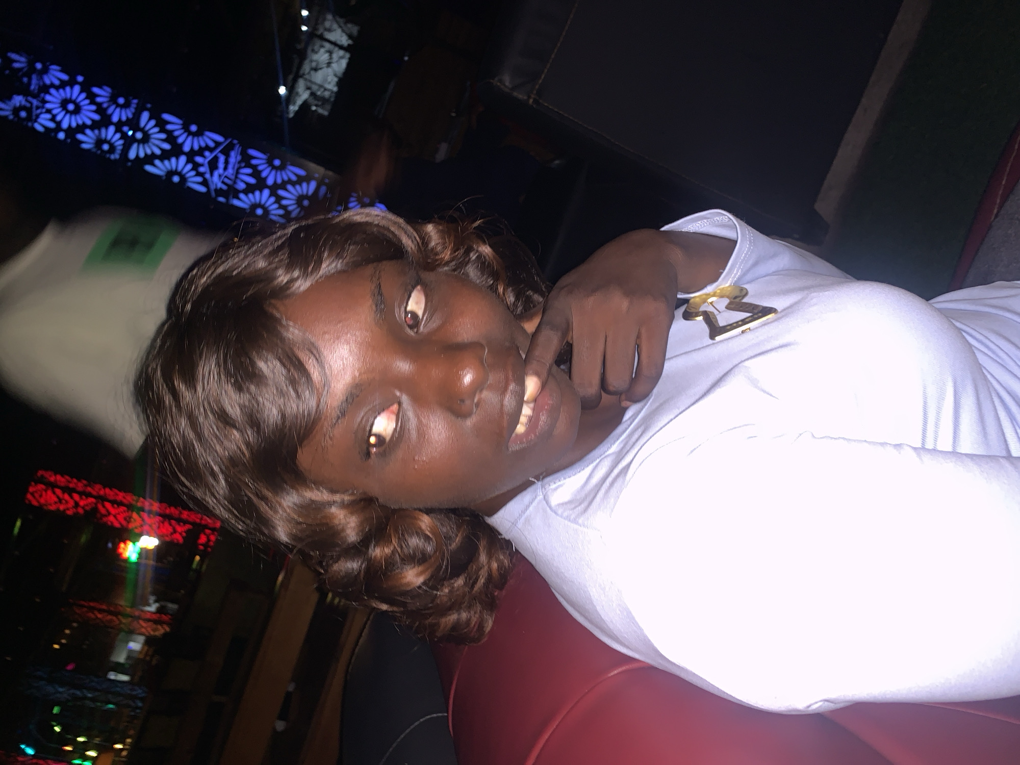 A dark-skinned woman with curly brown hair is seated in a red booth at night. She is wearing a light blue long-sleeved shirt with a gold pendant on it. She is looking to her right with her hand on her chin, her mouth slightly open. The background is dark with colorful lights, suggesting a public venue such as a restaurant or club. The image appears to be taken in a dimly lit environment, possibly Howayi, Nigeria, given the context.