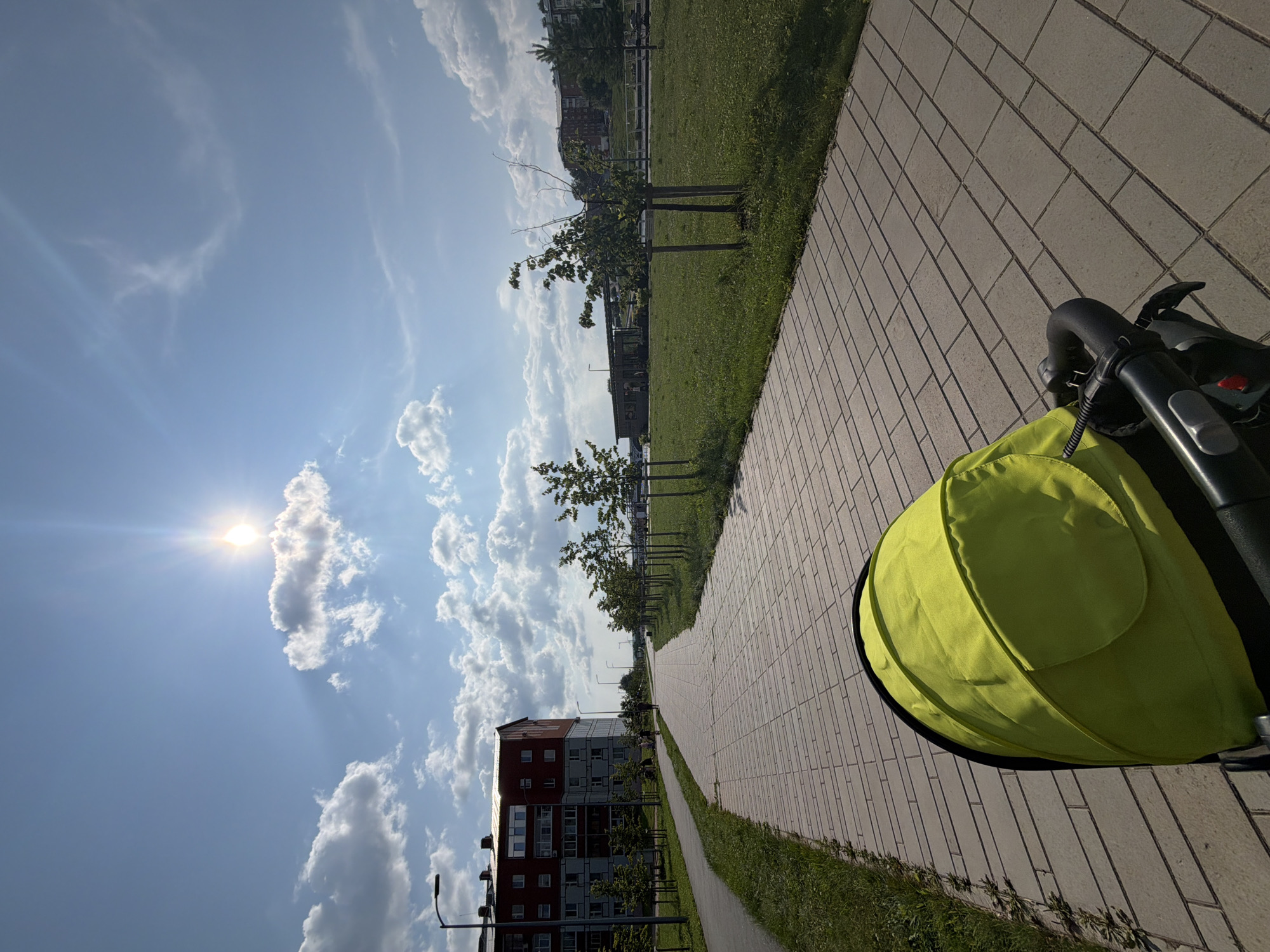 The image depicts a sunny day in Votkinsk, Russia, with a perspective from a stroller. In the foreground, the bright yellow canopy of a stroller is visible, with its dark grey handle and some control buttons. The stroller is on a paved walkway made of rectangular grey bricks, laid in a staggered pattern. To the left of the walkway, a strip of green grass runs alongside it, bordered by more grass. Beyond the grass, a grey paved path is visible with a streetlamp. Further in the background, a multi-story red and beige apartment building stands on the left side of the image. Several small, young trees with wooden stakes are planted in a neat row on the right side of the walkway, in a well-maintained grassy area. In the distance, more buildings and trees are visible under a bright blue sky dotted with white, fluffy clouds. The sun is high in the sky, casting bright rays and lens flares. A few distant figures can be seen walking on the paths.
