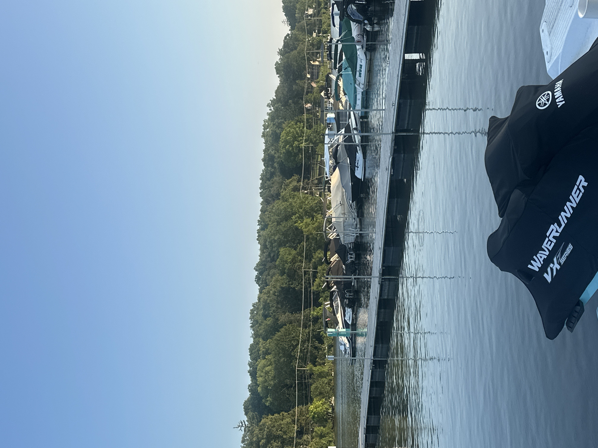 The image shows a marina on a lake in Algonquin, Illinois, USA. The sky is clear and blue. In the foreground, a jet ski cover with "Waverunner VX Series" and a Yamaha logo is visible. A dock extends into the water, with several boats moored alongside it. The boats are of various sizes and types, with some covered by tarps. In the background, a lush green treeline forms the shore of the lake, with some houses and power lines visible through the trees. The water is calm and reflects the sky and the surrounding scenery.