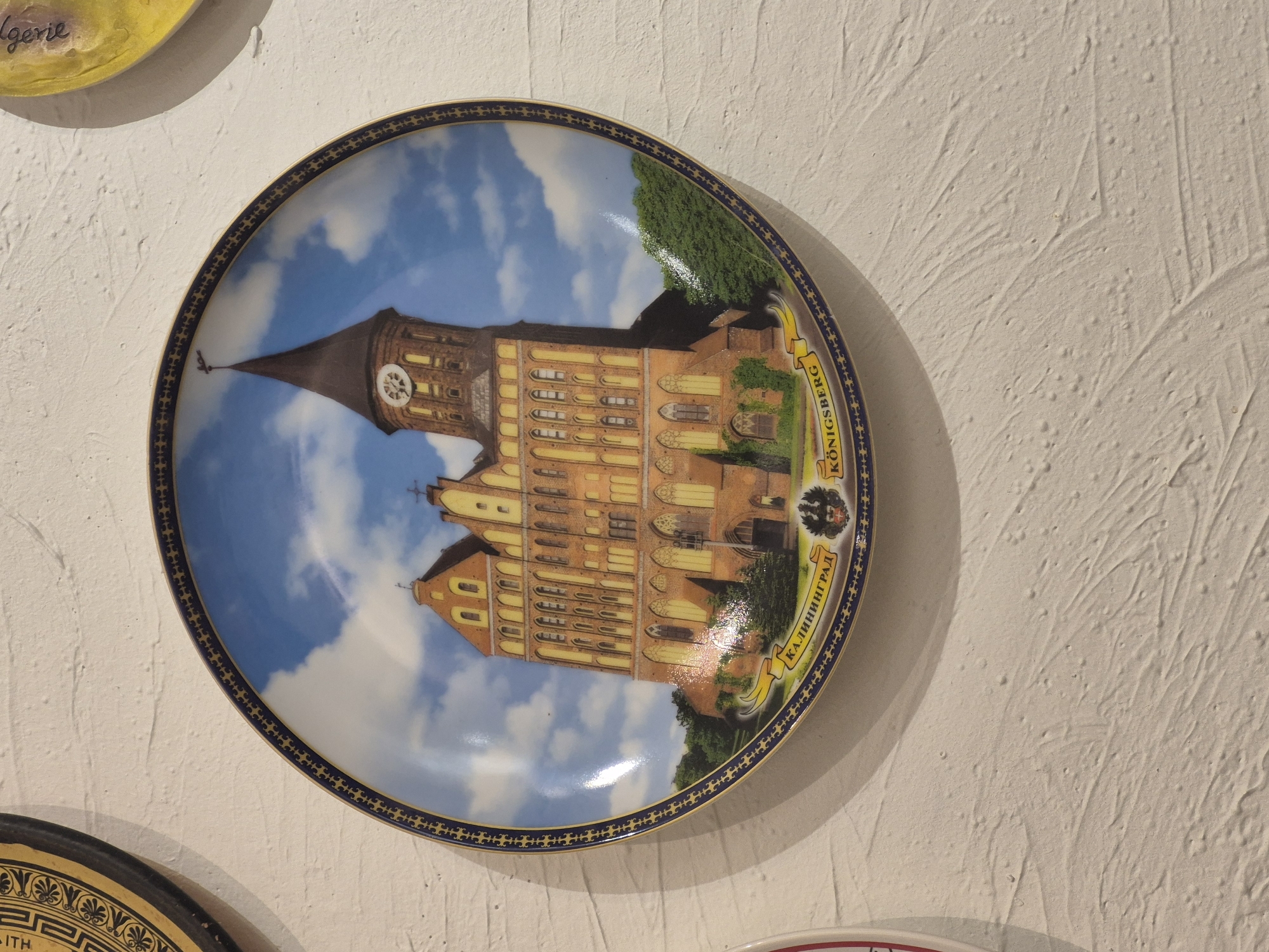 The image shows a decorative plate featuring a depiction of a large, ornate brick building with a tall, pointed spire and a clock tower. The building has many windows and architectural details characteristic of gothic or neo-gothic styles. The plate has a decorative blue and gold border with a repeating pattern. A banner beneath the building displays text in two languages: "КА.ЛИНИНГРАД" (Kaliningrad) and "KÖNIGSBERG". The plate is mounted on a textured white wall. There are no people or animals visible in the image. The location in Krasnodar, Russia is not determinable from the image itself.